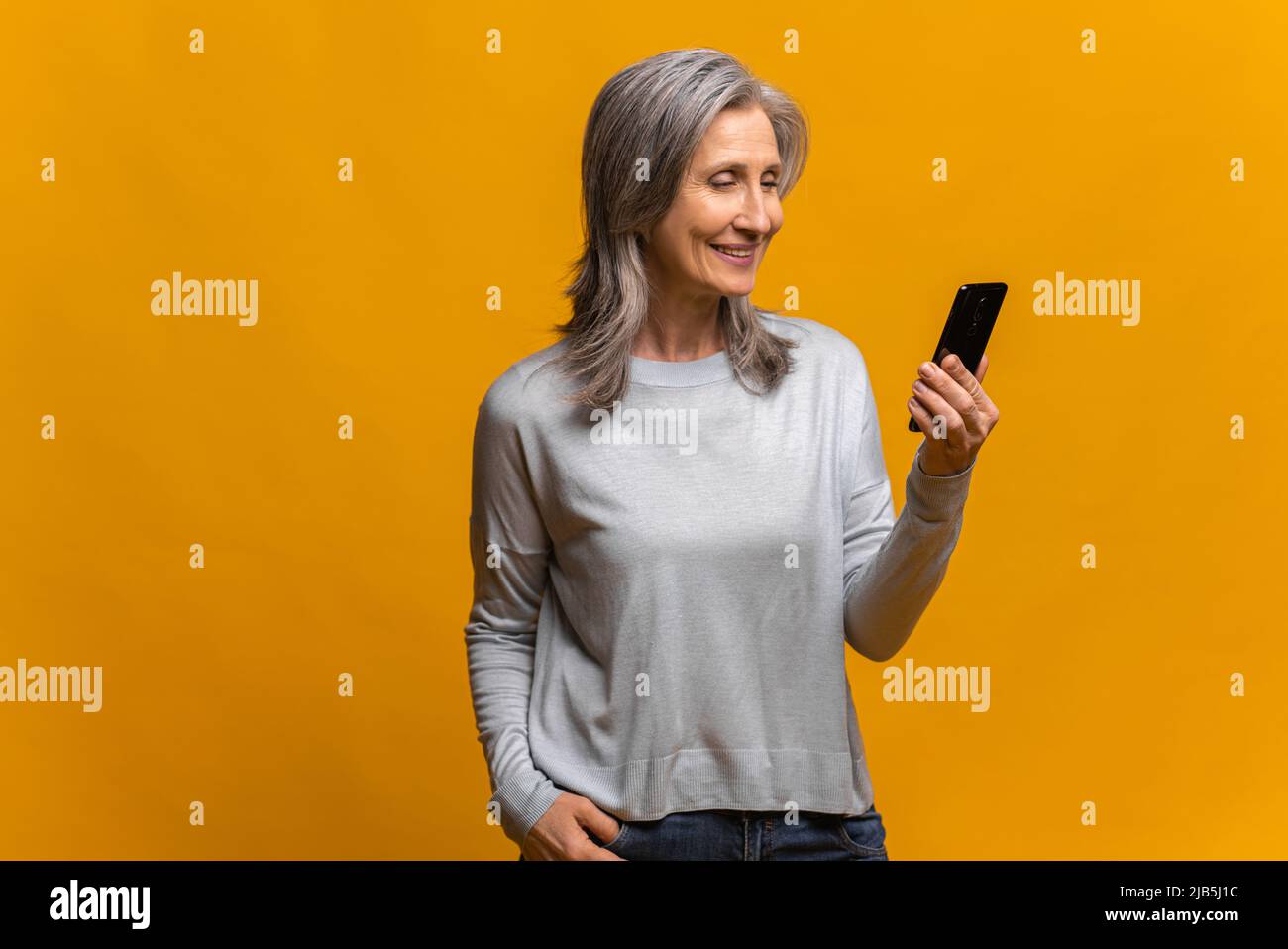 Profilportrait der glücklichen trendigen reifen Dame mit Smartphone und lächelnd, zufrieden mit der Verbindungsqualität. Innenaufnahme des Studios isoliert auf gelbem Hintergrund Stockfoto