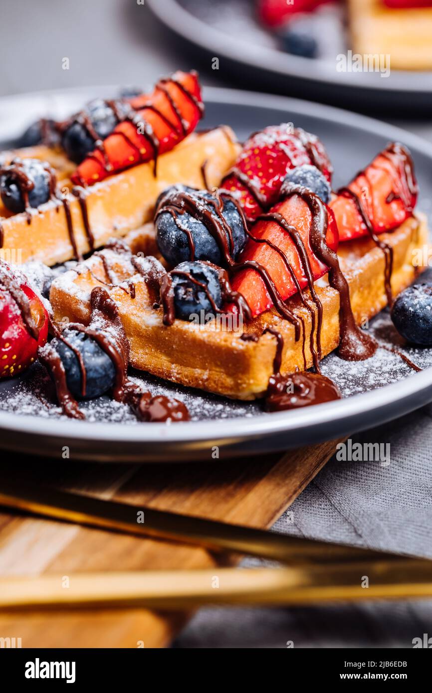 Belgische Waffel mit Schokolade, Erdbeere, Heidelbeeren und Puderzucker auf dunklem Teller Stockfoto