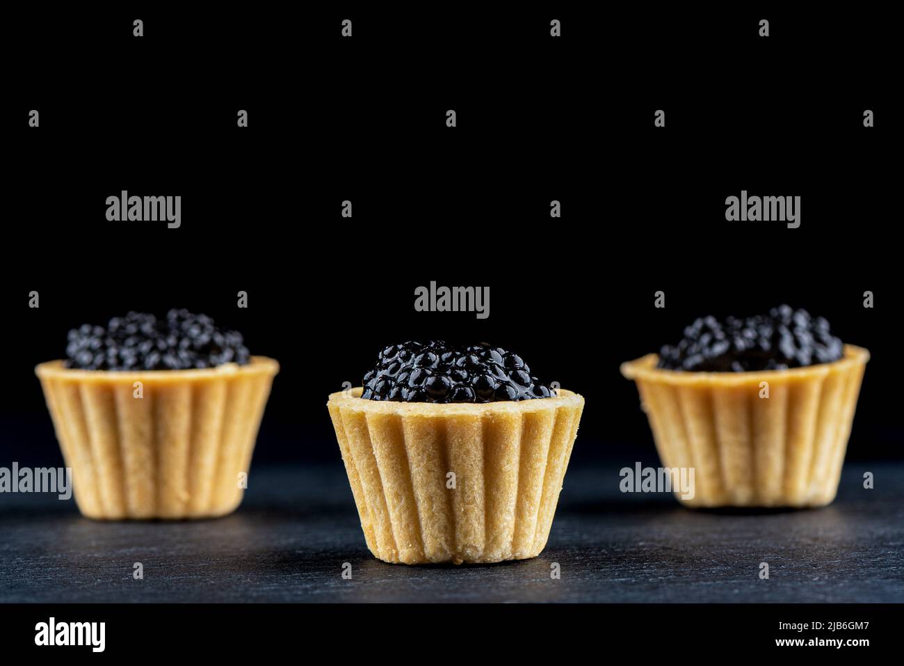 Selektiver Fokus auf den vorderen Tart mit schwarzem Kaviar. Drei Törtchen mit schwarzem Kaviar auf dunklem Grund. Die hinteren Torten sind verschwommen. Stockfoto