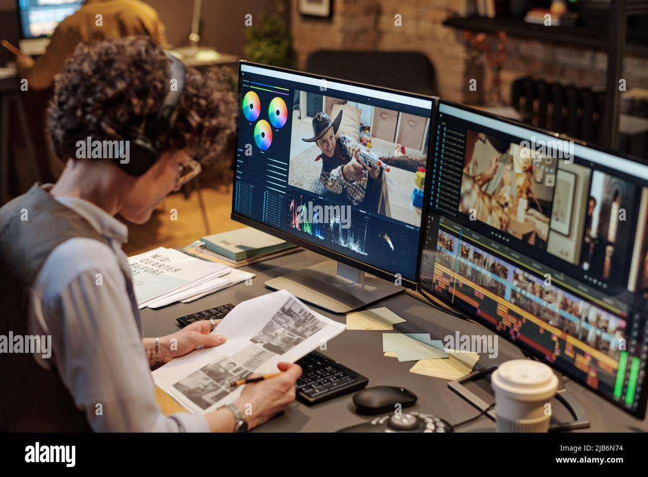 Junge Redakteurin untersucht Skript, während sie am Tisch am Computer mit großem Display arbeitet Stockfoto