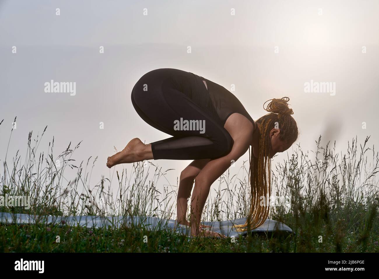 Sportliche Frau mit geflochtener Frisur, die beim aktiven Training an der frischen Luft auf den Händen balanciert. Sportliche weibliche Person, die während des erstaunlichen Sonnenaufgangs im Sommer trainiert. Stockfoto