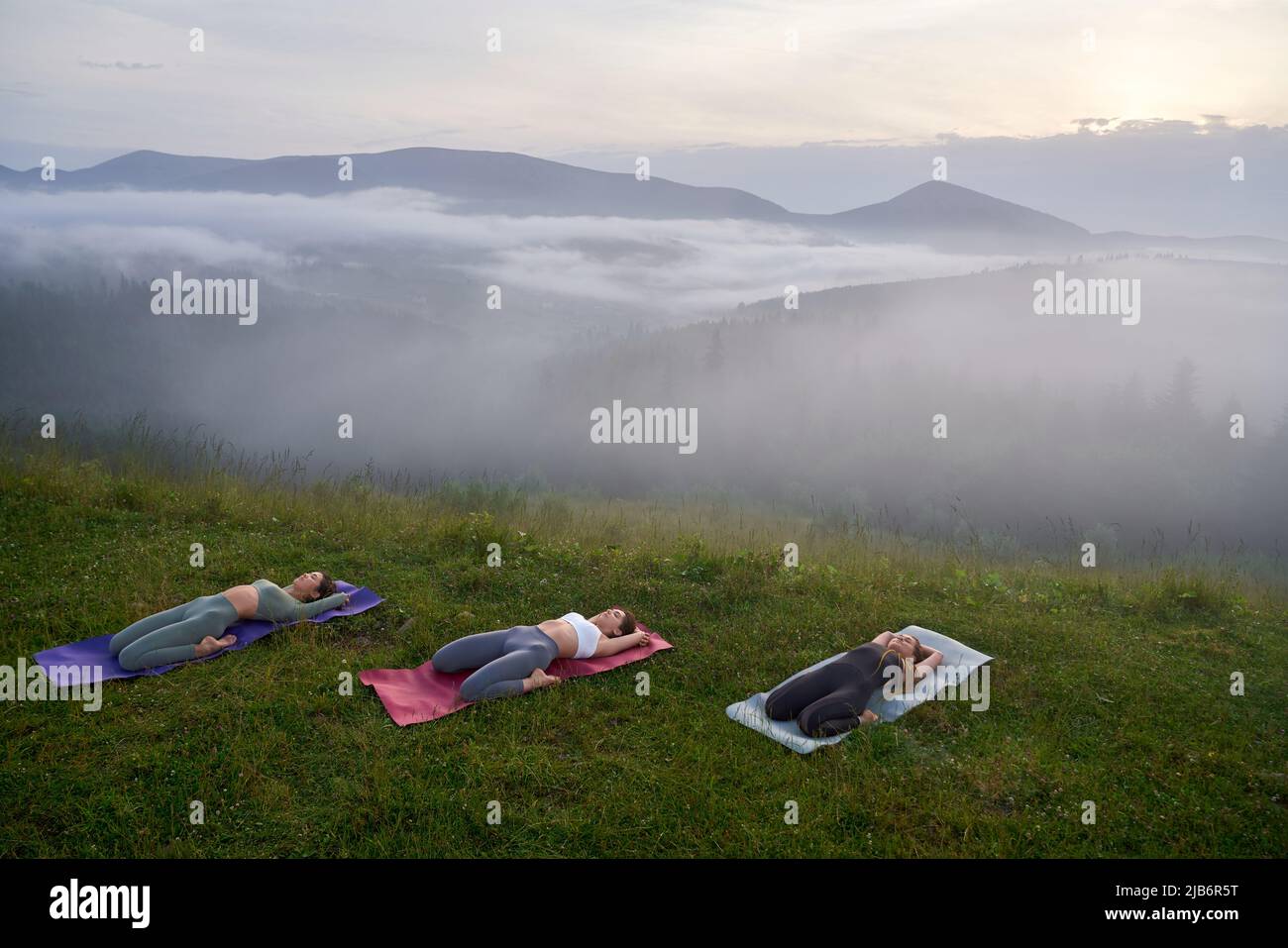 Drei junge Frauen in Sportkleidung liegen auf einer Yogamatte, während sie während des Trainings den Körper strecken. Aktive Frauen, die in der Freizeit im Freien trainieren. Stockfoto
