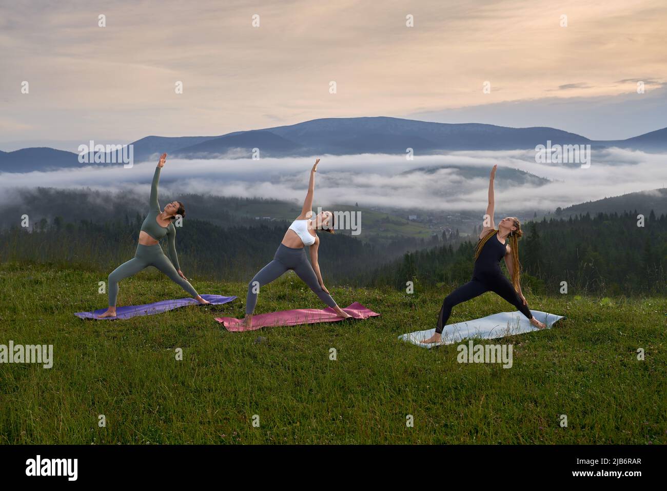 Junge sportliche Frauen in Sportkleidung üben Yoga mit Sommerbergen auf dem Hintergrund. Gruppe von drei Frauen, die an der frischen Luft aktiv trainieren. Stockfoto