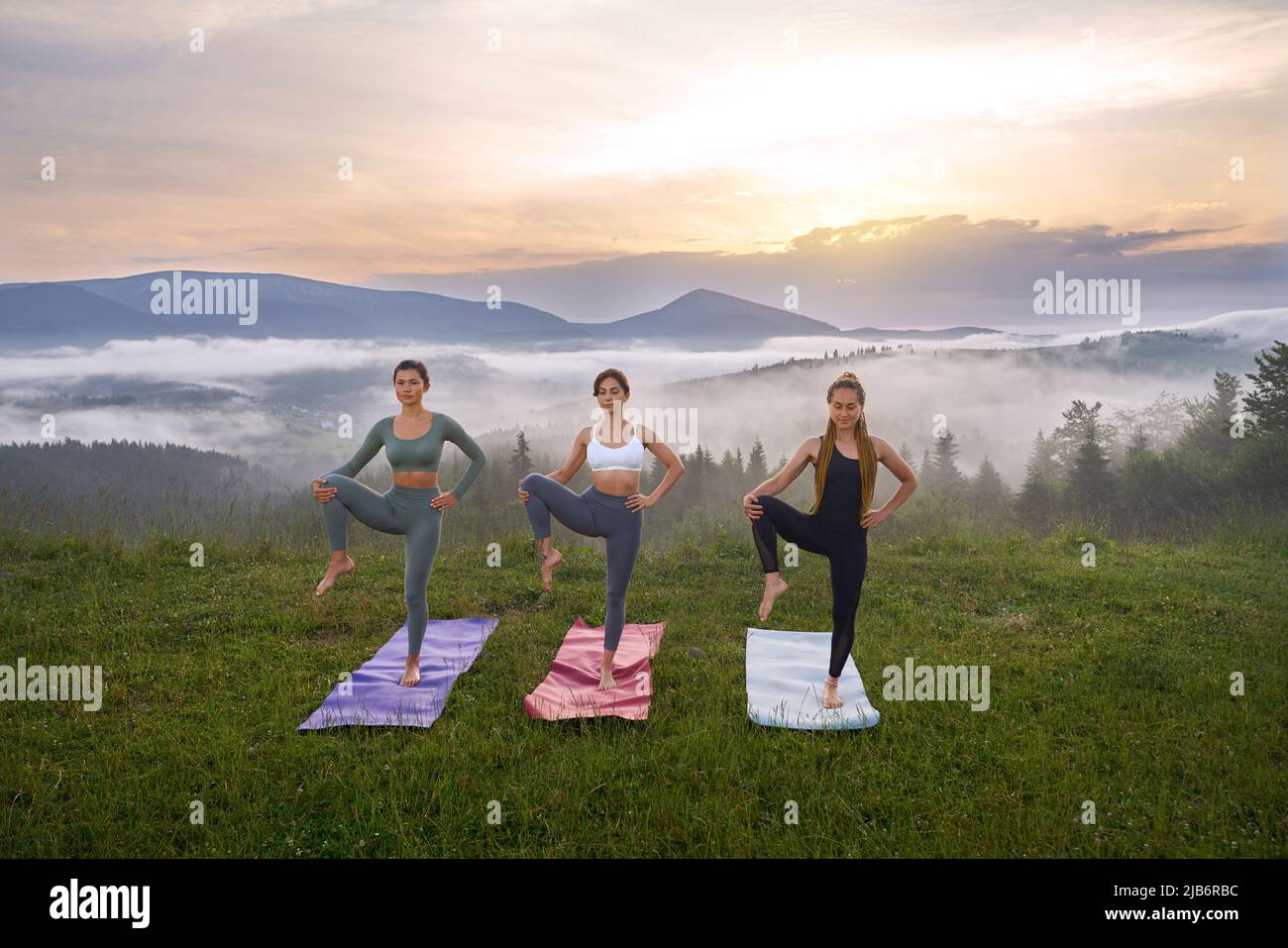 Eine Gruppe von drei jungen Frauen in Sportkleidung, die Zeit für Yoga an der frischen Luft verbringen. Schöner Sommeruntergang im Hintergrund. Stockfoto