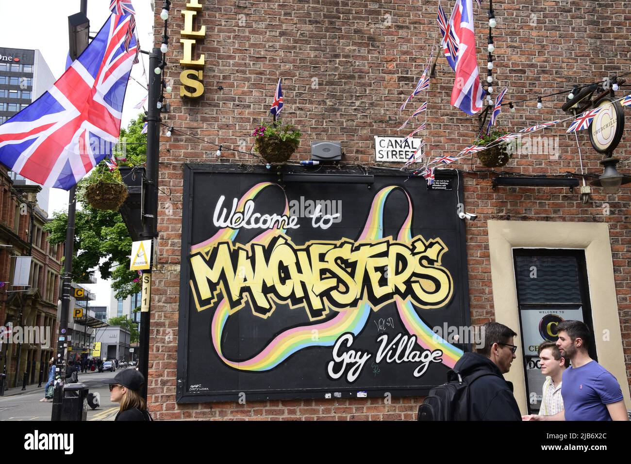 Manchester, Großbritannien, 3.. Juni 2022. Die Menschen laufen an einem Schild „Welcome to Manchester's Gay Village“, Canal Street, Manchester, Großbritannien, vorbei. Union Jack Flaggen, um das Jubiläum zu feiern. Tägliches Leben auf den Straßen von Manchester, England, Großbritannien und den Britischen Inseln am zusätzlichen Jubilee Bank Holiday Freitag. Am Freitag, dem 3.. Juni, ist der offizielle Bankfeiertag des Platinum Jubilee, ein zusätzlicher Bankfeiertag zur Feier des 70.-jährigen Bestehens der Königin als Monarch. Quelle: Terry Waller/Alamy Live News Stockfoto