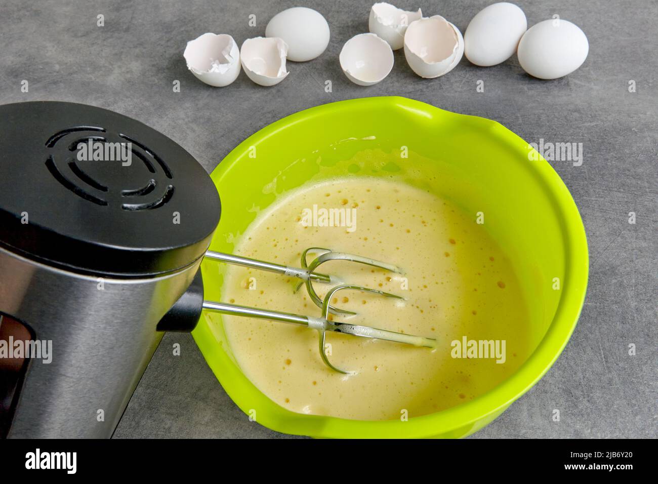 Eier mit Zucker in einer grünen Schüssel mit einem in einen Mixer eingetauchten Quirl und Eierschalen aus Hühnereiern auf einem Küchentisch aus dunklem Beton geschlagen. bize wird gemacht Stockfoto