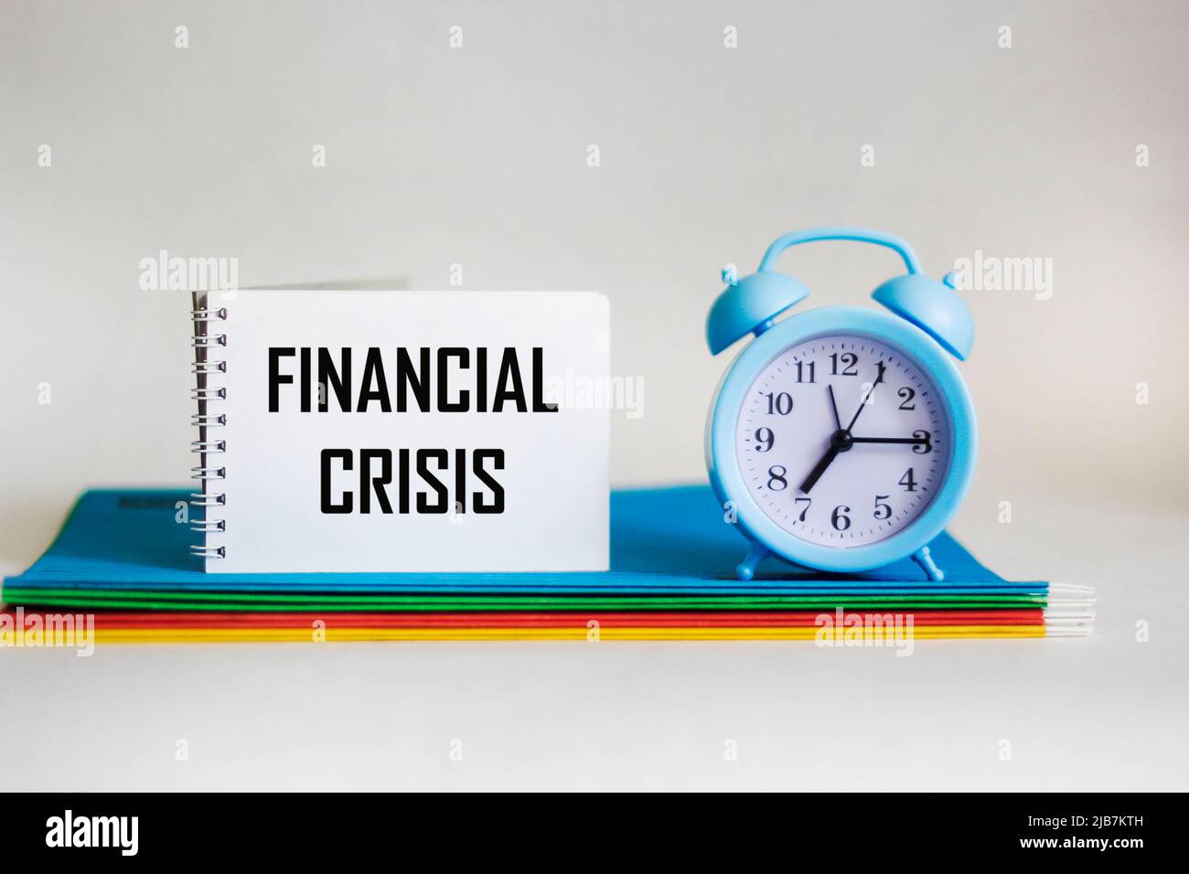 Das Wort Finanzkrise steht auf einem Notizblock, der auf mehrfarbigen Mappen und neben einer Uhr auf weißem Hintergrund liegt. Stockfoto