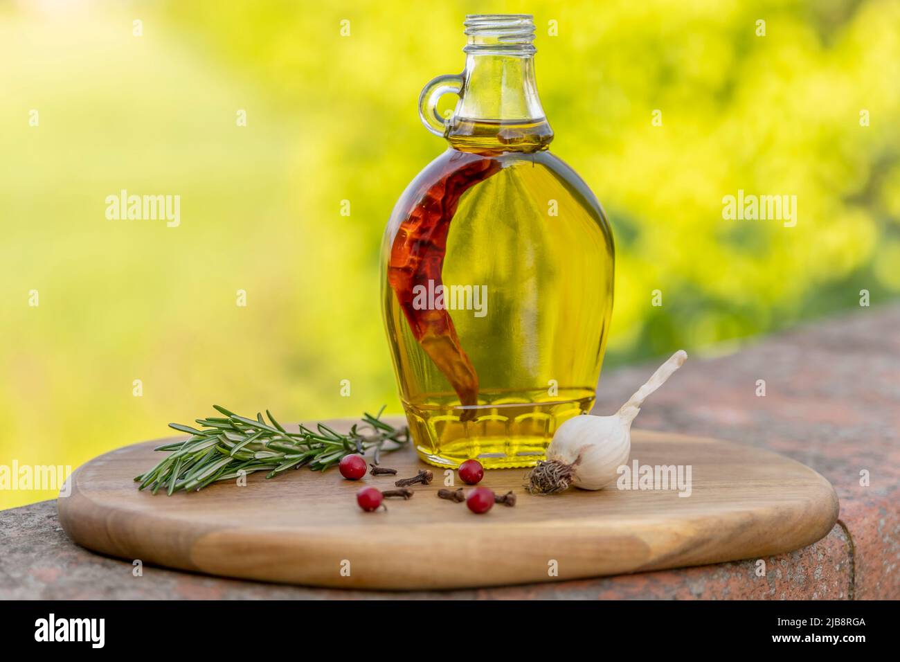 Auf einem hölzernen Schneidebrett mit Knoblauch und Rosmarin befindet sich ein Glas mit extra nativem Olivenöl, das mit rotem Chili gewürzt ist Stockfoto