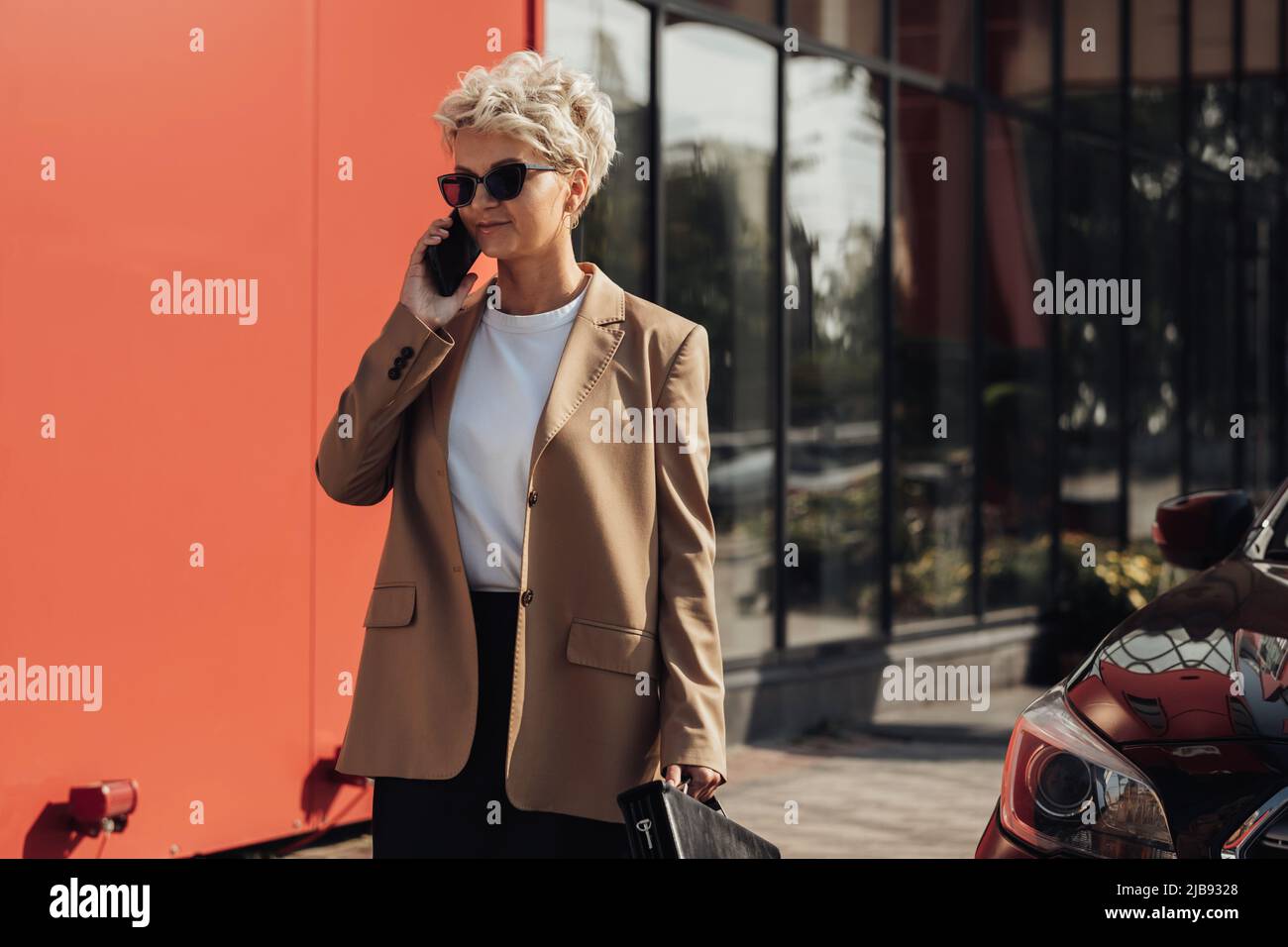 Modische Geschäftsfrau mit Aktenkoffer in der Hand im Telefongespräch in der Nähe des Luxuswagens Stockfoto