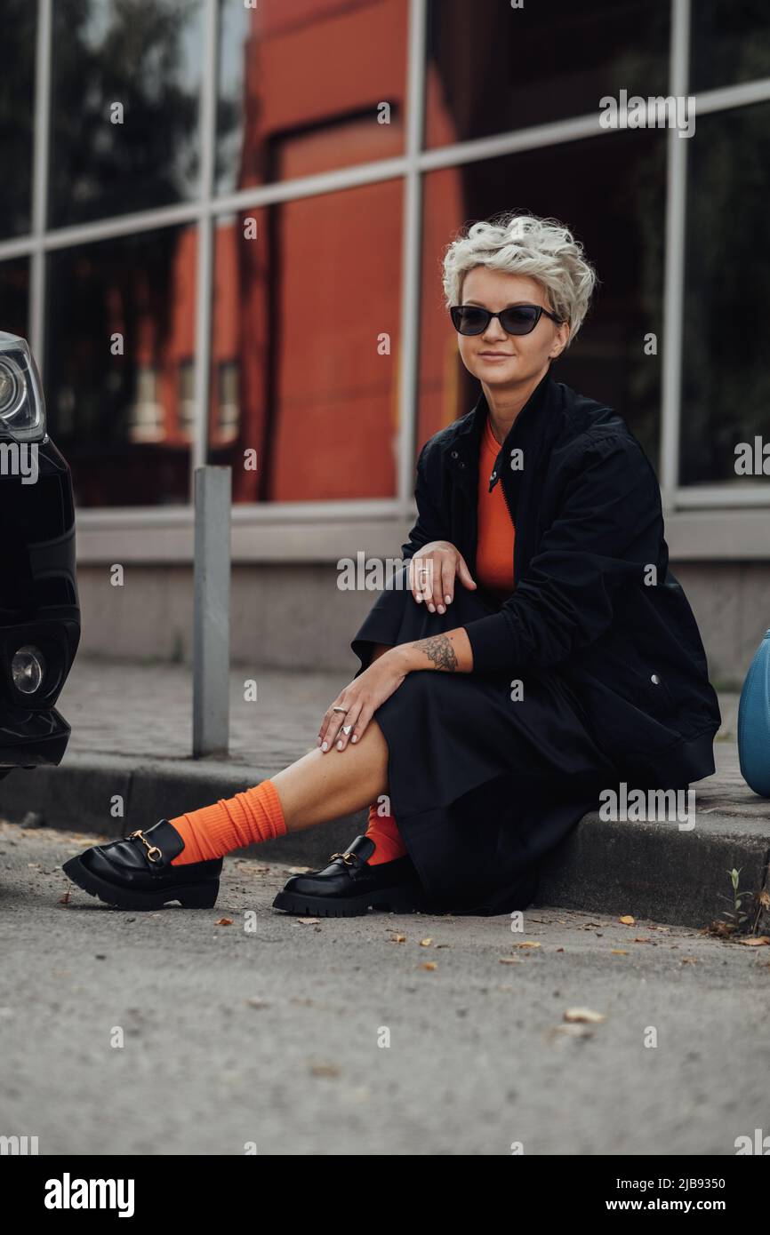 Stilvolle junge Frau in Jacke und Sonnenbrille auf dem Boden in der Nähe des Autos sitzen Stockfoto