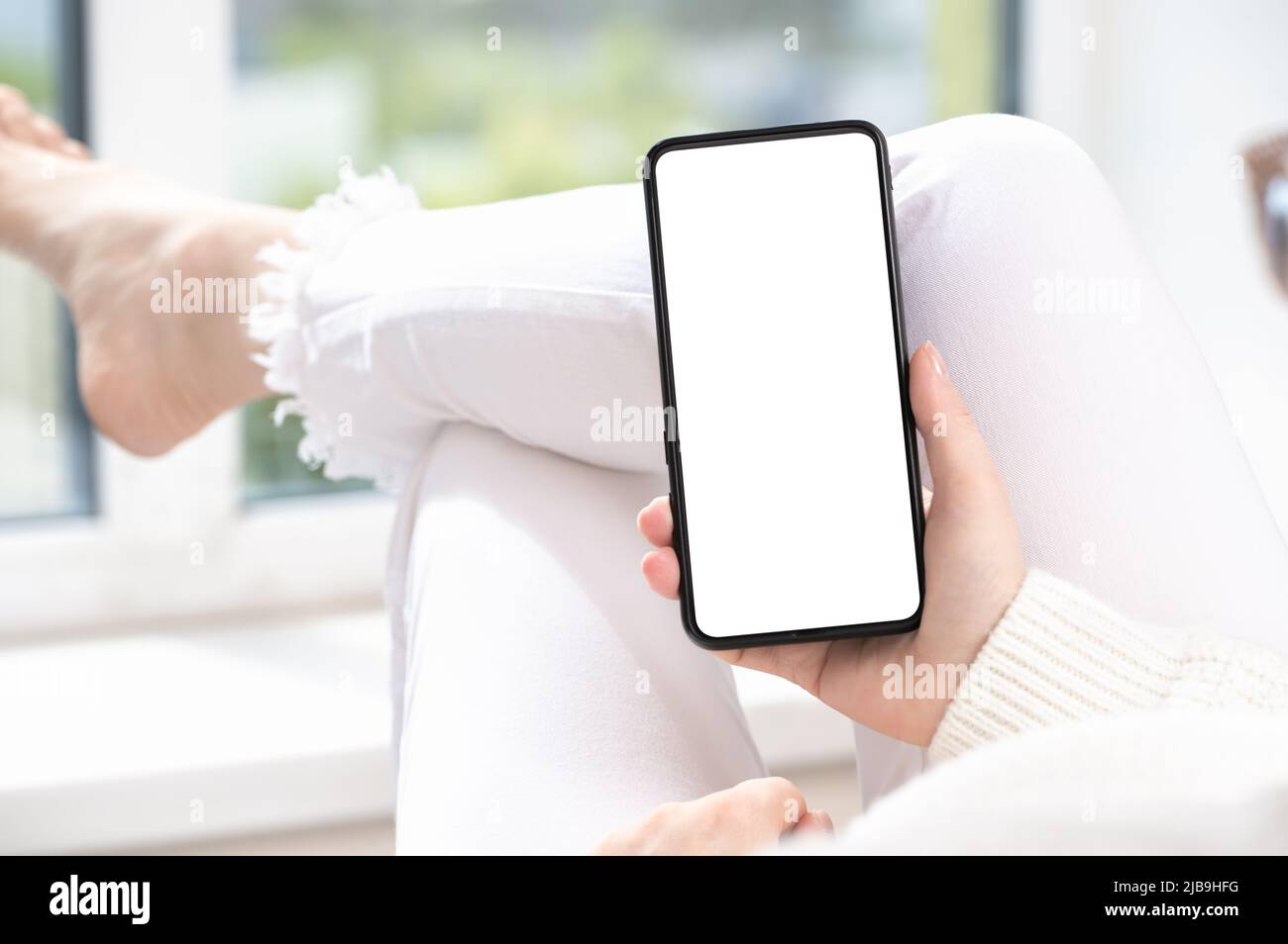Frau in weißer Kleidung liegt zu Hause mit gekreuzten Beinen am Fenster und nutzt Handy oder Smartphone. Nutzen Sie die mobile Shopping-App, schauen Sie sich Social-Media-Nachrichten an, oder lesen Sie Text Stockfoto