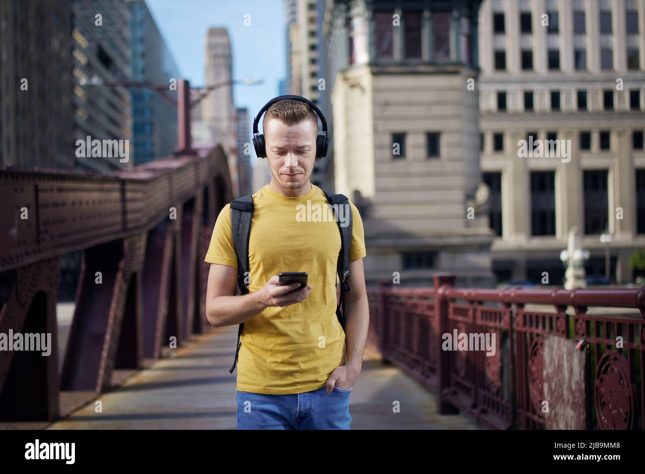 Junger Mann, der mit kabellosen Kopfhörern Musik hört und während eines Spaziergangs durch die Stadt in sein Mobiltelefon blickt. Stockfoto