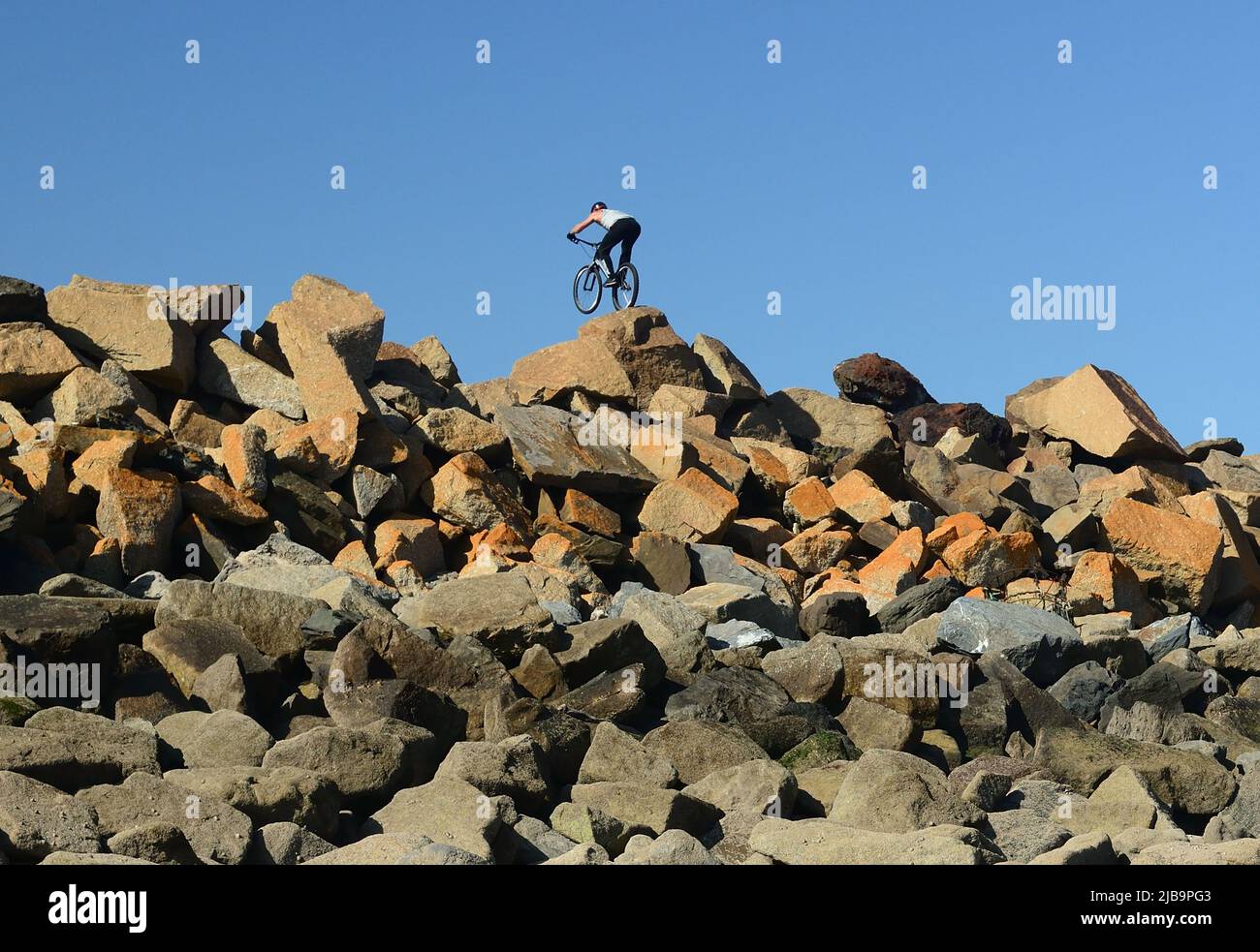 Ein akrobatischer Radfahrer, der Stunts auf mehrfarbigen Felsenpanzern auf der Meeresabwehr ausführt, vom Strand unten bei Ebbe gesehen. Stockfoto