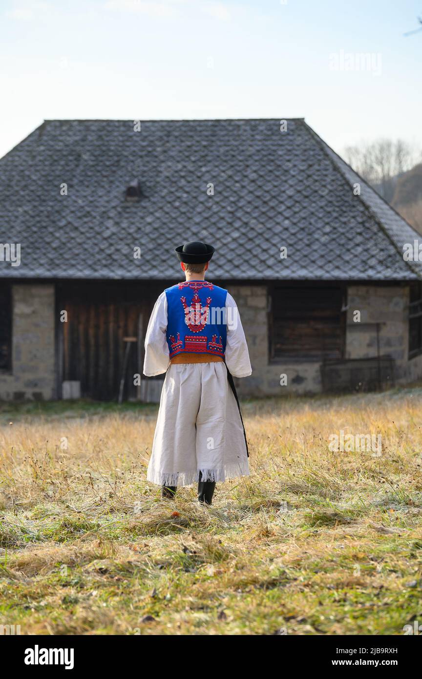 Ein Mann in traditioneller Tracht. Slowakische Kostüm im Herbst Natur. Altes Landhaus im Hintergrund. Details des slowakischen Kostüms von De Stockfoto