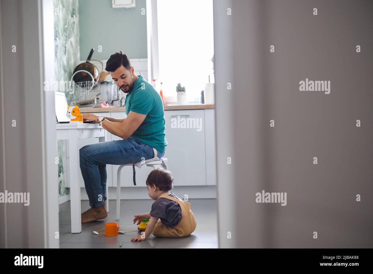 Vater arbeitet am Laptop, während das Kind auf dem Küchenboden spielt Stockfoto