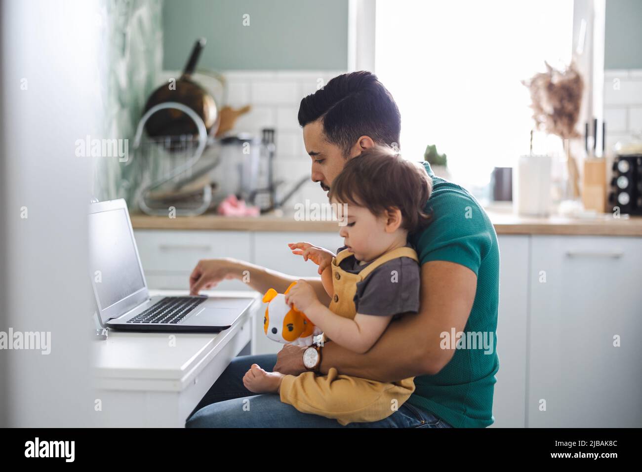 Vater arbeitet am Laptop, Kind sitzt auf seinem Schoß Stockfoto