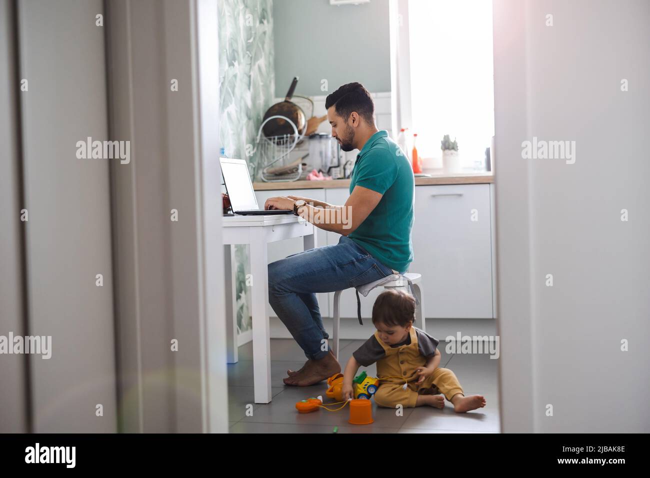 Vater arbeitet am Laptop, während das Kind auf dem Küchenboden spielt Stockfoto