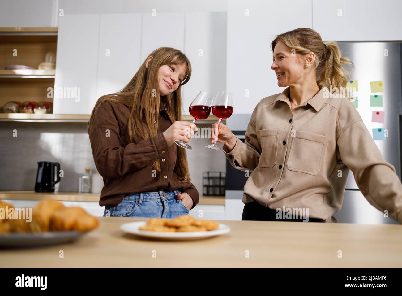 Zwei Frauen unterschiedlichen Alters feiern in der Küche etwas. Eine hübsche Frau mittleren Alters und ihre Erwachsene Tochter trinken Wein und plaudern bei ho Stockfoto