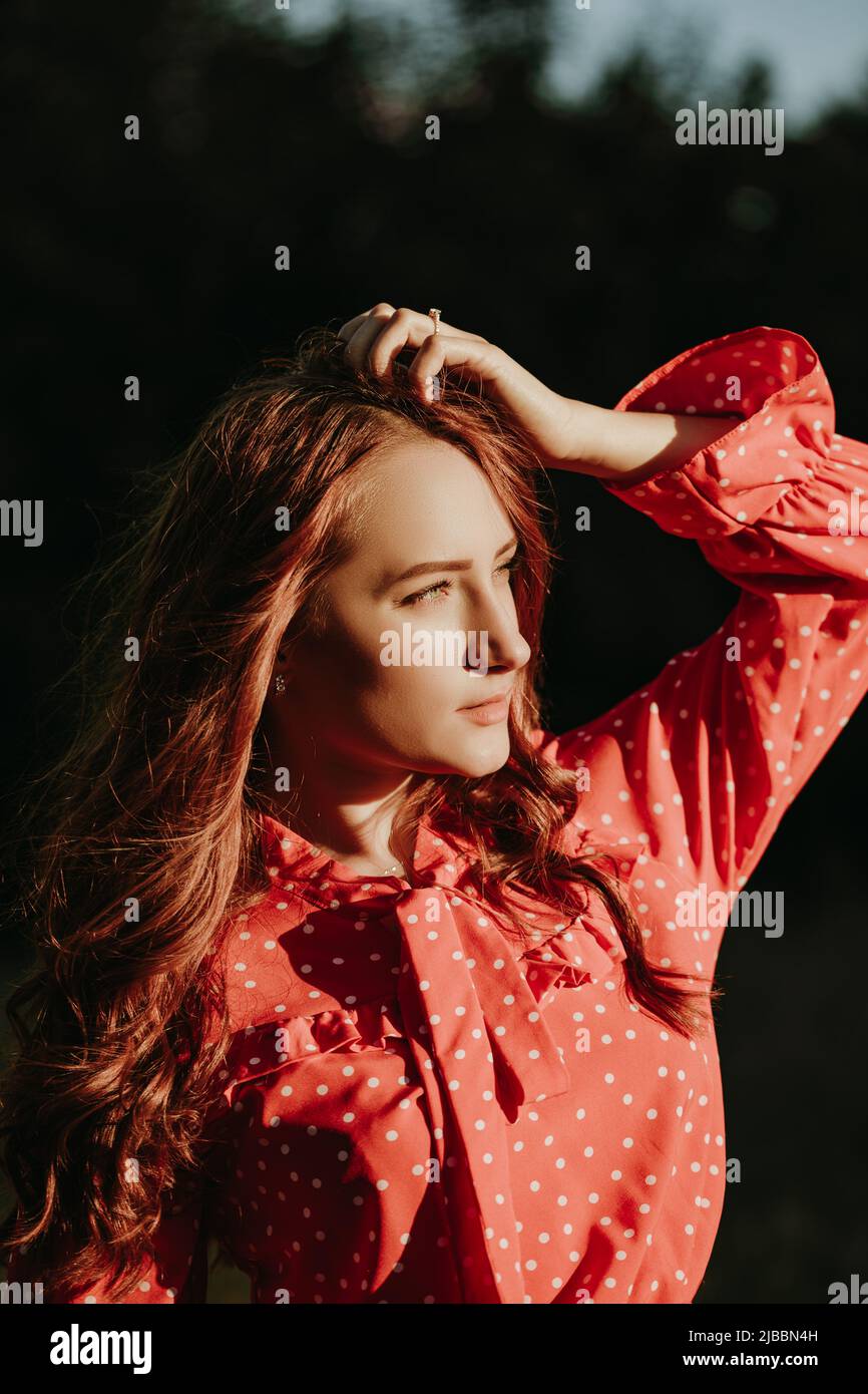 Portait der Schönheit junge rothaarige Frau mit langen lockigen dicken Haaren in rotem Hemdkleid mit weißen Punkten posiert auf einer Kamera im Sonnenlicht gekleidet Stockfoto