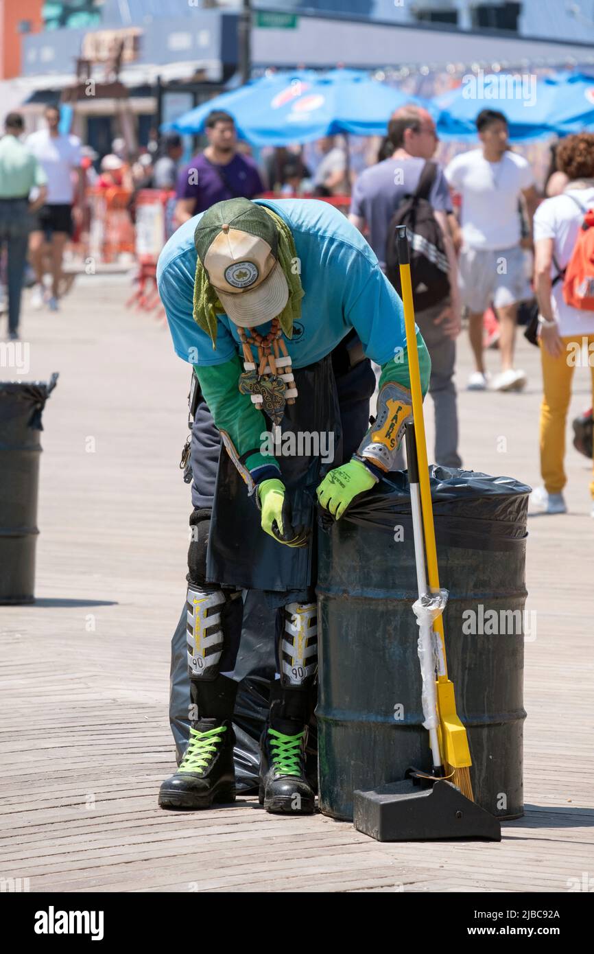 Ein anonymer Mitarbeiter der New Yorker Parkabteilung mit viel Schutzausrüstung und seinem eigenen Stilbewusstsein wechselt Plastikmüllbeutel auf der Promenade von Coney Island. Stockfoto