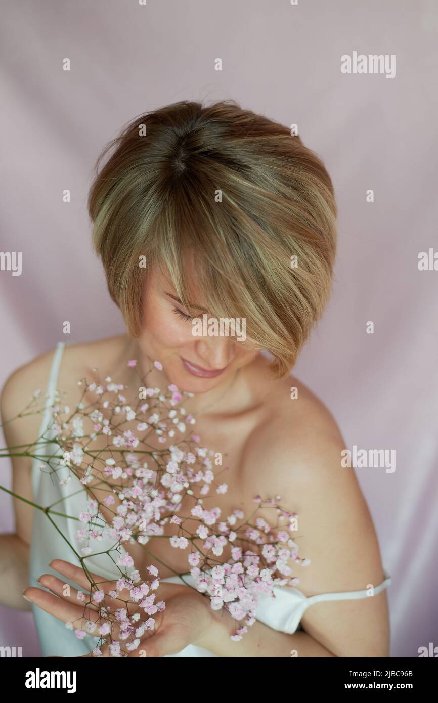 Porträt eines Mädchens auf einem rosa Hintergrund mit Blumen. Zärtlichkeit und feminin. Die Sphäre der Schönheit und der Gesundheit der Frauen. Kurzer Haarschnitt, blondes Haar Stockfoto