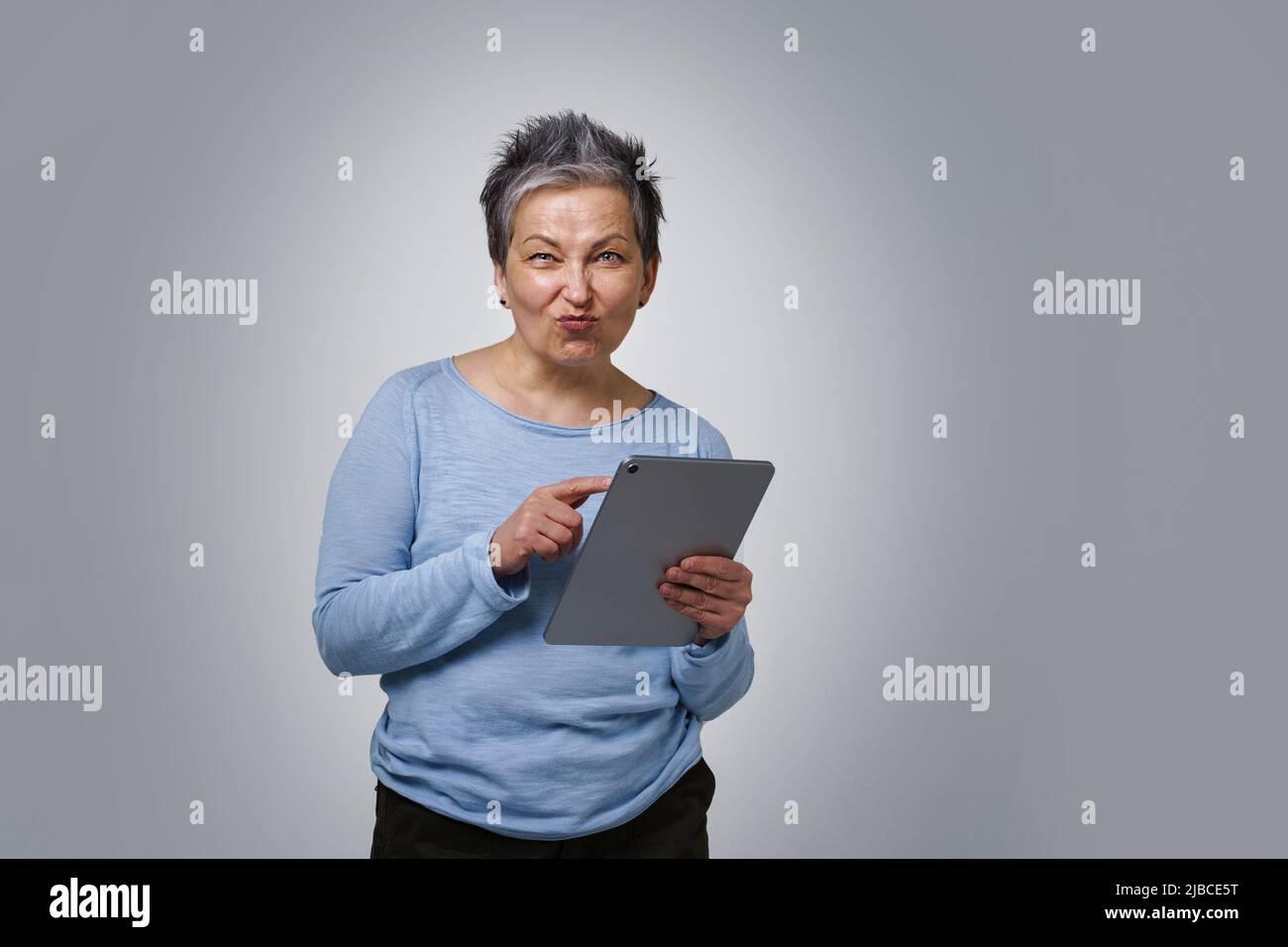 Verspielte grauhaarige reife Frau mit digitalem Tablet, die in sozialen Medien arbeitet oder nachcheckt. Hübsche Frau im Jahr 50s in blauer Bluse isoliert auf weißem Hintergrund. Ältere Menschen und Technologien. Stockfoto
