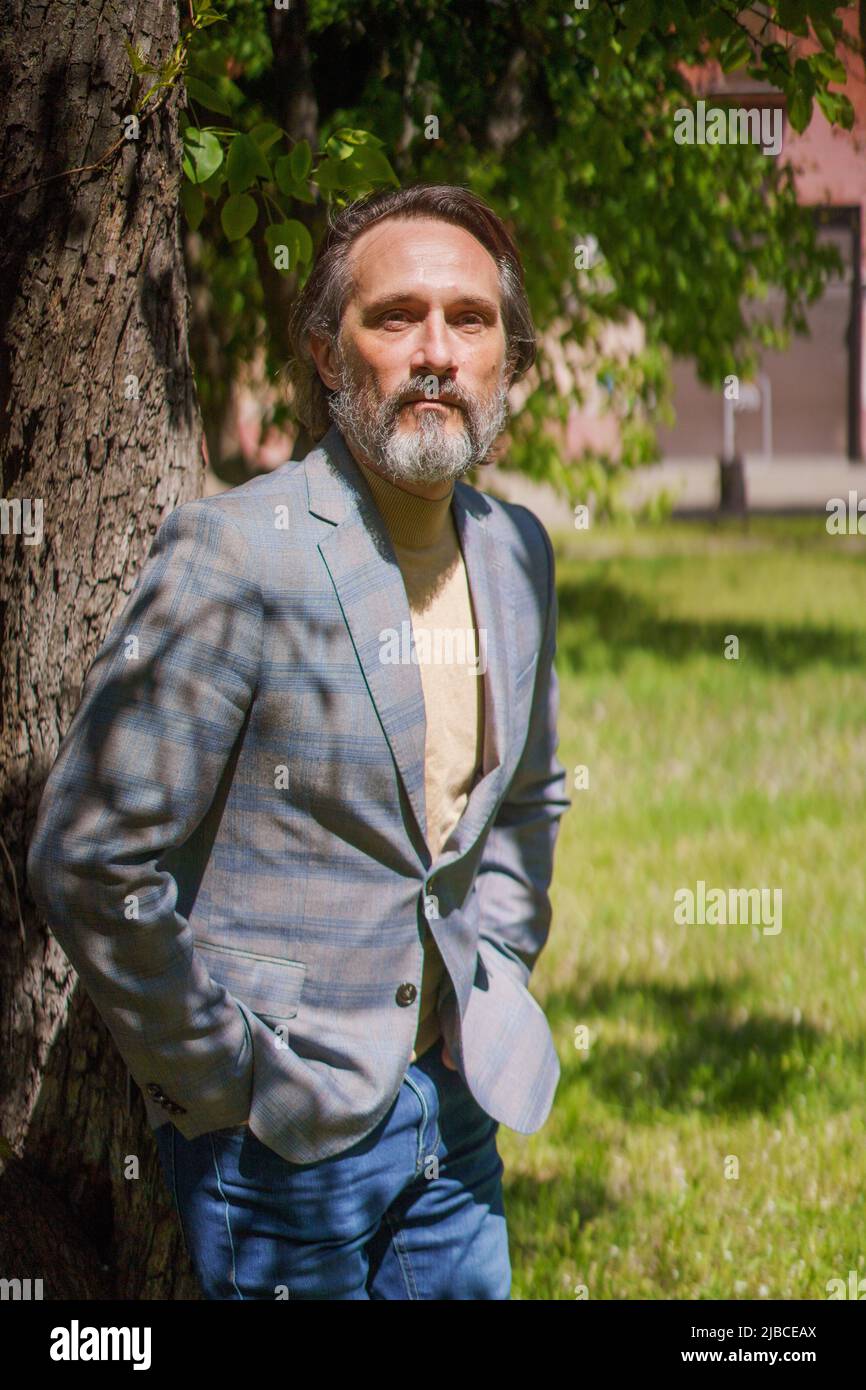 Lehnte sich an einen hübschen Mann mit grauem Bart und Blick auf die Kamera in einer lässigen grauen Jacke und einer blauen Jeans. Leben nach 40 Jahren Konzept, Probleme und Depressionen. Krise des mittleren Alters. Stockfoto