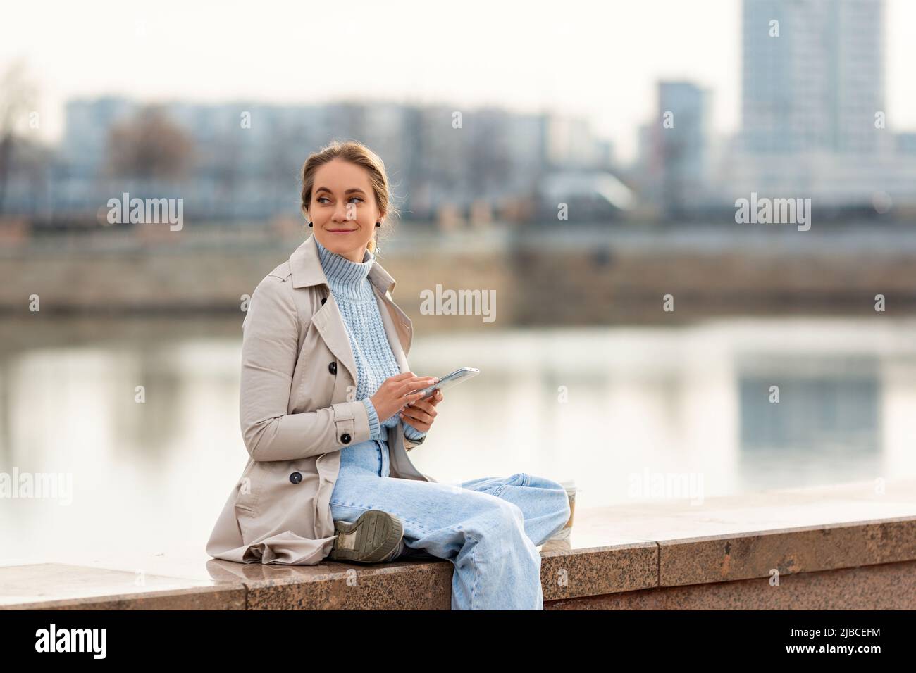 Eine junge Frau mit Smartphone an einem warmen Frühlingstag. Sie lächelt und schaut weg. Stockfoto