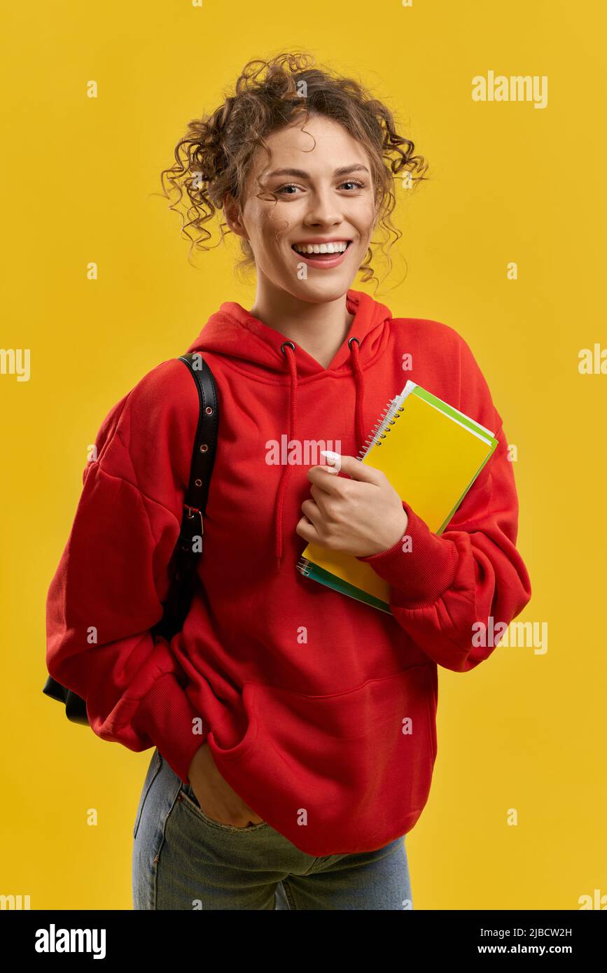 Nettes Mädchen mit Rucksack, Blick auf die Kamera, während Notizen in der Halle. Vorderansicht einer Studentin, die mit der Hand in der Tasche stand, lächelnd, isoliert auf gelbem Studiohintergrund. Konzept des Studierens. Stockfoto