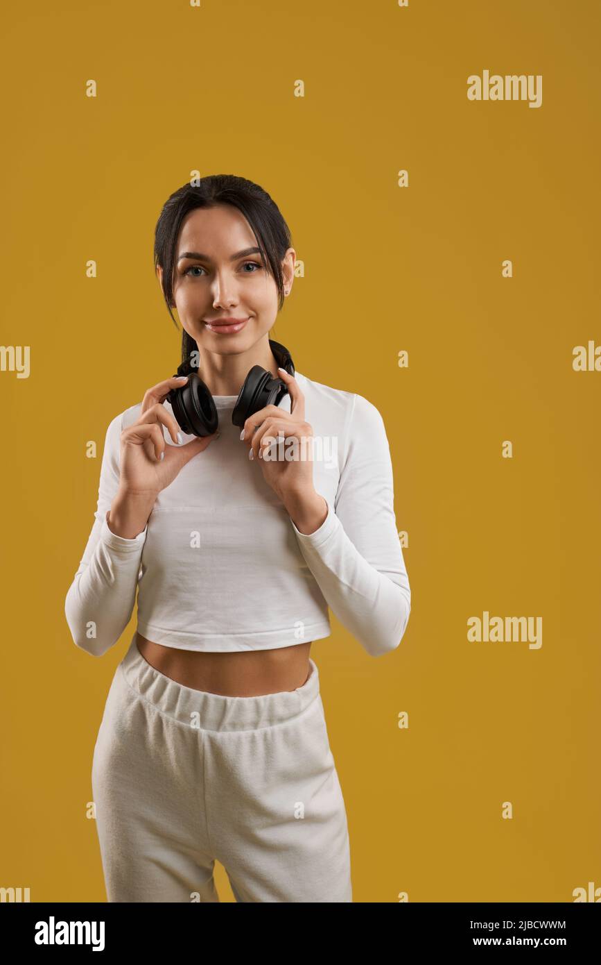 Kaukasische Frau in sportlichem Outfit mit Headset, die im Studio auf die Kamera schaut. Porträtansicht einer lächelnden Frau mit kabellosen Kopfhörern auf dem Nackentraining, isoliert auf orange. Konzept des Lebensstils. Stockfoto