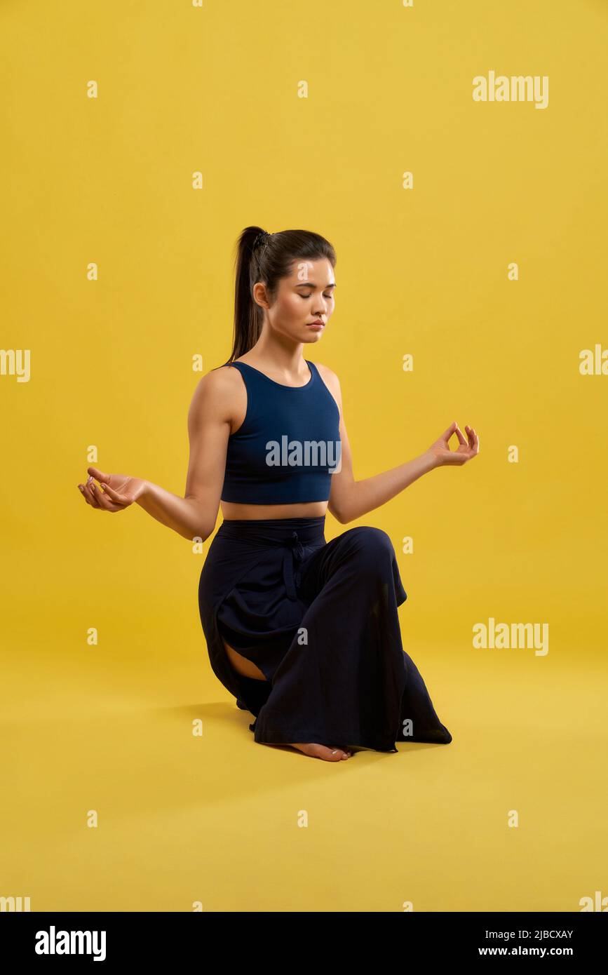 Ruhige weibliche Hockstellung mit Mudra-Händen und Yoga-Übungen in der Halle. Vorderansicht eines jungen Mädchens in schwarzer Spitze, meditierend, isoliert auf gelbem Hintergrund, Kopierraum. Konzept der Meditation. Stockfoto