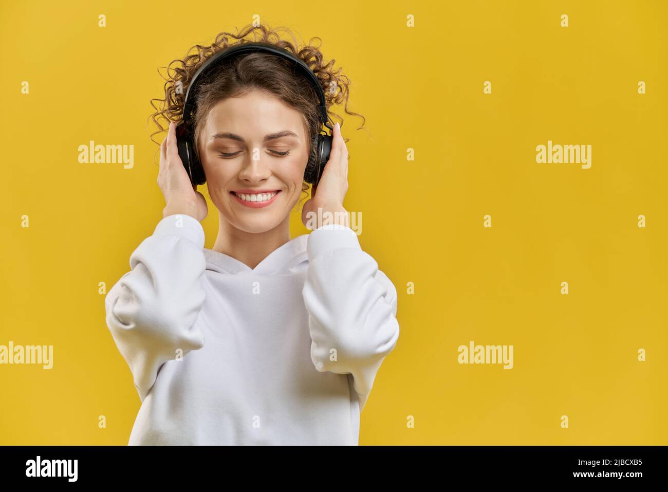 Fröhliche Frau mit modernen Ohrhörern, die Musik hören, Spaß in der Halle haben. Porträtansicht von Musikliebhaberinnen, die Sound genießen, mit geschlossenen Augen, isoliert auf orangefarbenem Studiohintergrund. Konzept des Genusses. Stockfoto