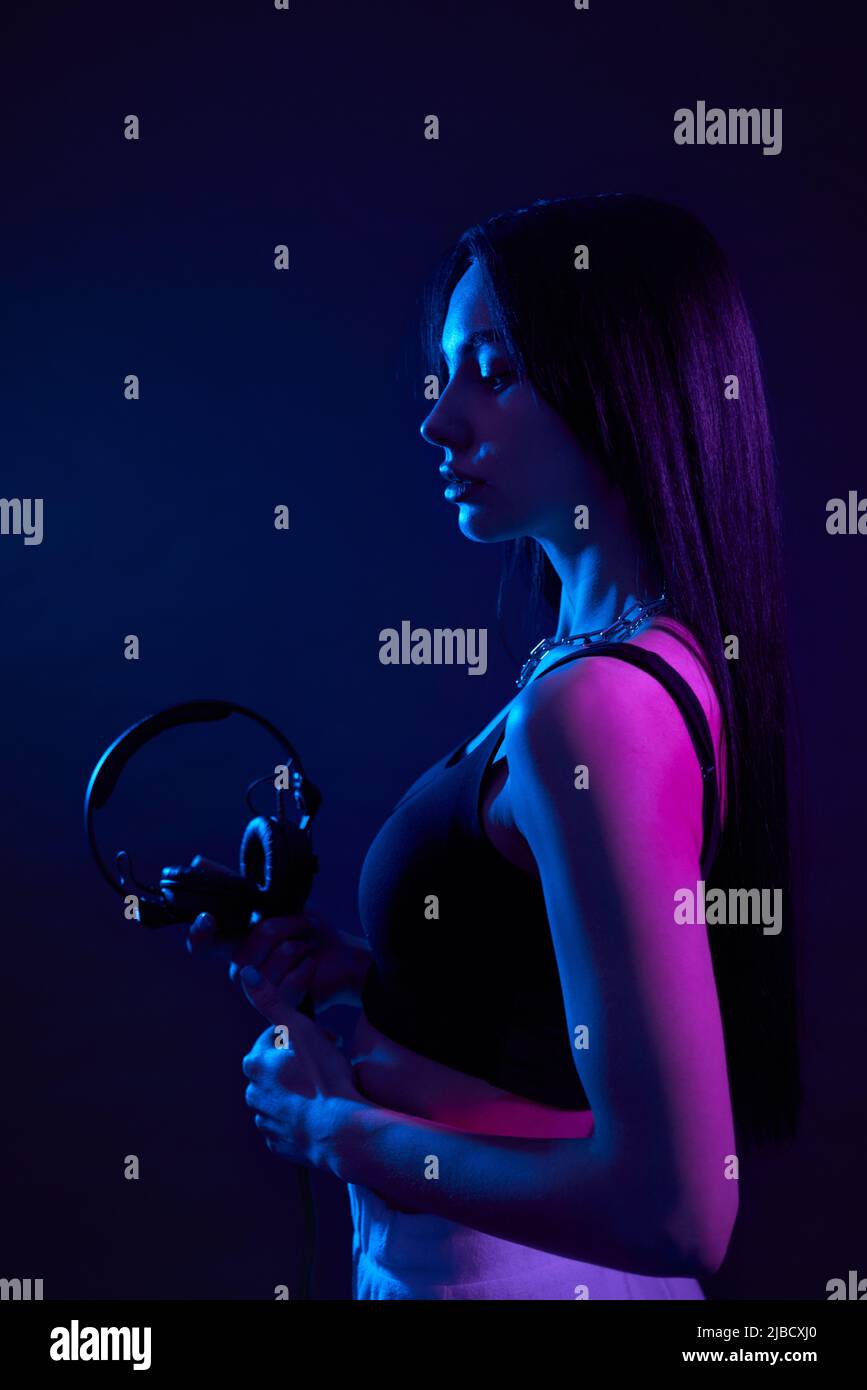 Anmutiges Mädchen mit schwarzen Ohrhörern, im Studio mit violettem Licht beleuchtet. Seitenansicht einer ruhigen Dame mit geradem, dunklem Haar, das ein großes Headset hält, isoliert auf dunkelblauem Hintergrund. Konzept des Posierens. Stockfoto