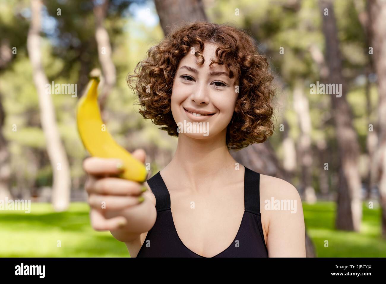 Junge Rotschopf-Frau in einem schwarzen Sport-BH, die auf dem Stadtpark steht, im Freien eine gesunde Banane hält und mit einem Smiley auf die Kamera schaut. Outdoo Stockfoto