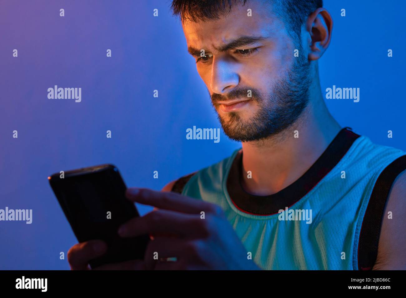 Porträt eines jungen kaukasischen Mannes mit Bart, der sein Handy benutzt. Sie wird mit blauen und orangefarbenen Lichtern beleuchtet. Stockfoto