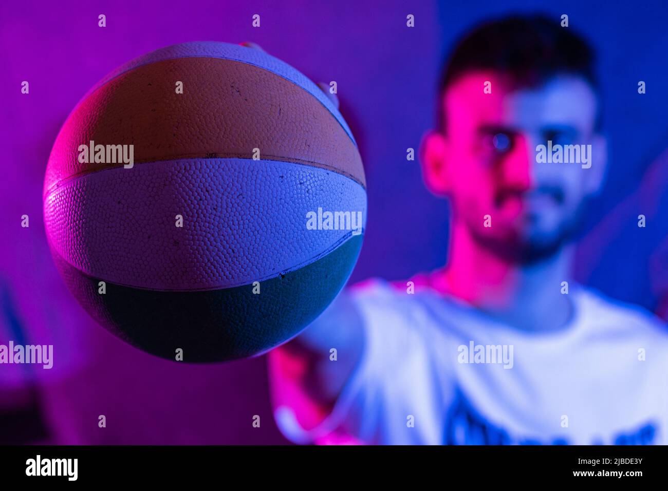 Ein junger kaukasischer bärtiger Mann mit ausgestrecktem Arm, der einen kleinen Basketball in verschiedenen Farben hält. Der Ball ist im Vordergrund und im Perso im Fokus Stockfoto