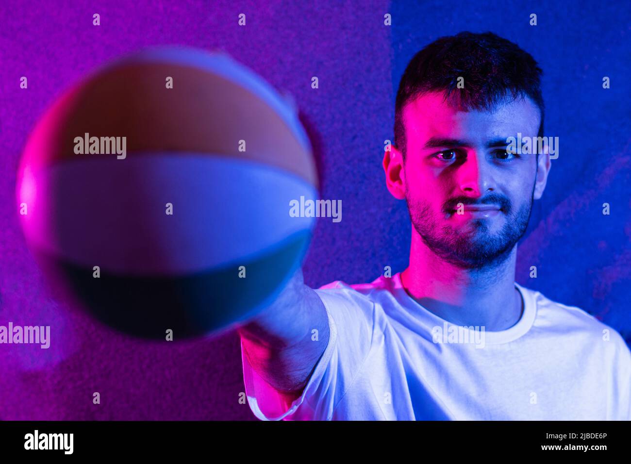 Ein junger kaukasischer bärtiger Mann mit ausgestrecktem Arm, der einen kleinen Basketball in verschiedenen Farben hält. Der Ball ist im Vordergrund nicht fokussiert und das p Stockfoto