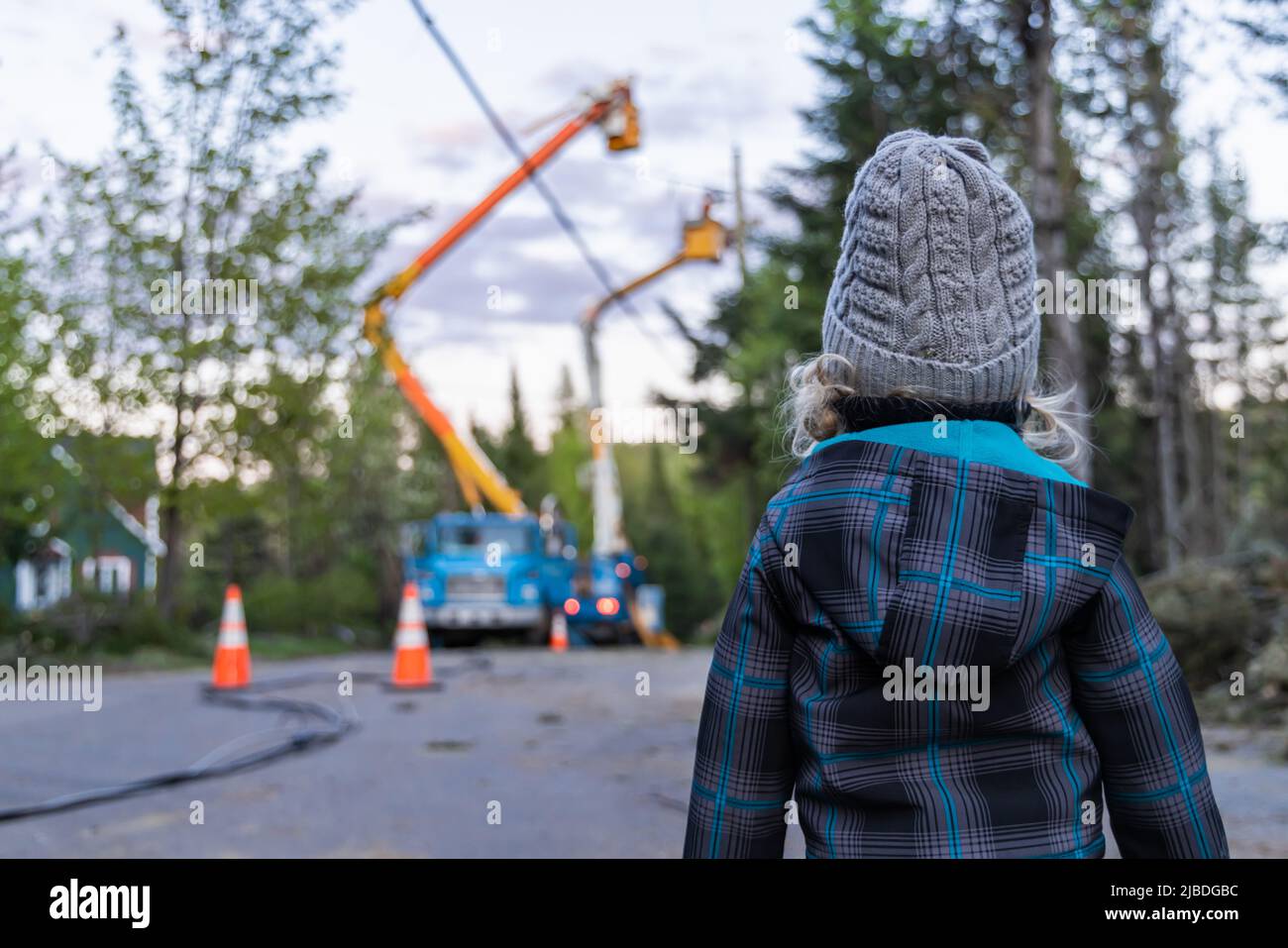 Rückansicht eines neugierigen, vierjährigen Jungen, der draußen steht, während die Powerline-Ingenieure im Hintergrund verschwommen sind, um den elektrischen Betrieb nach einem Sturm wiederherzustellen. Stockfoto