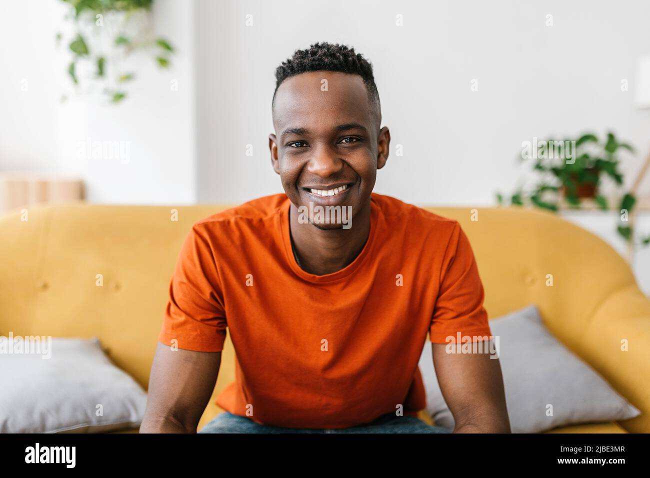Selbstbewusster junger erwachsener afrikanischer Mann, der zu Hause auf dem Sofa sitzt und die Kamera lächelt Stockfoto