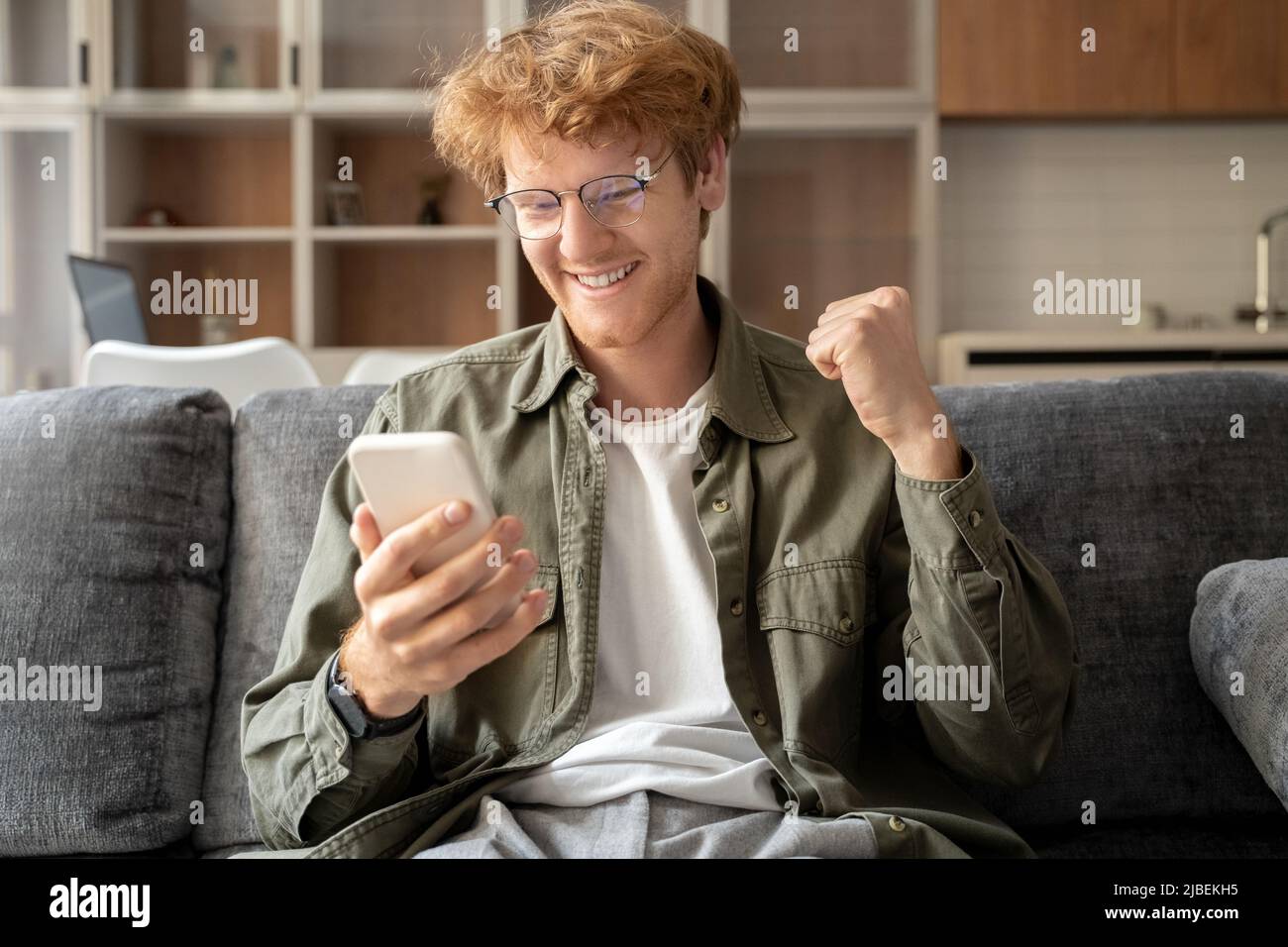 Junger britischer Ingwer in Brille hält Smartphone zeigt ja Geste Stockfoto