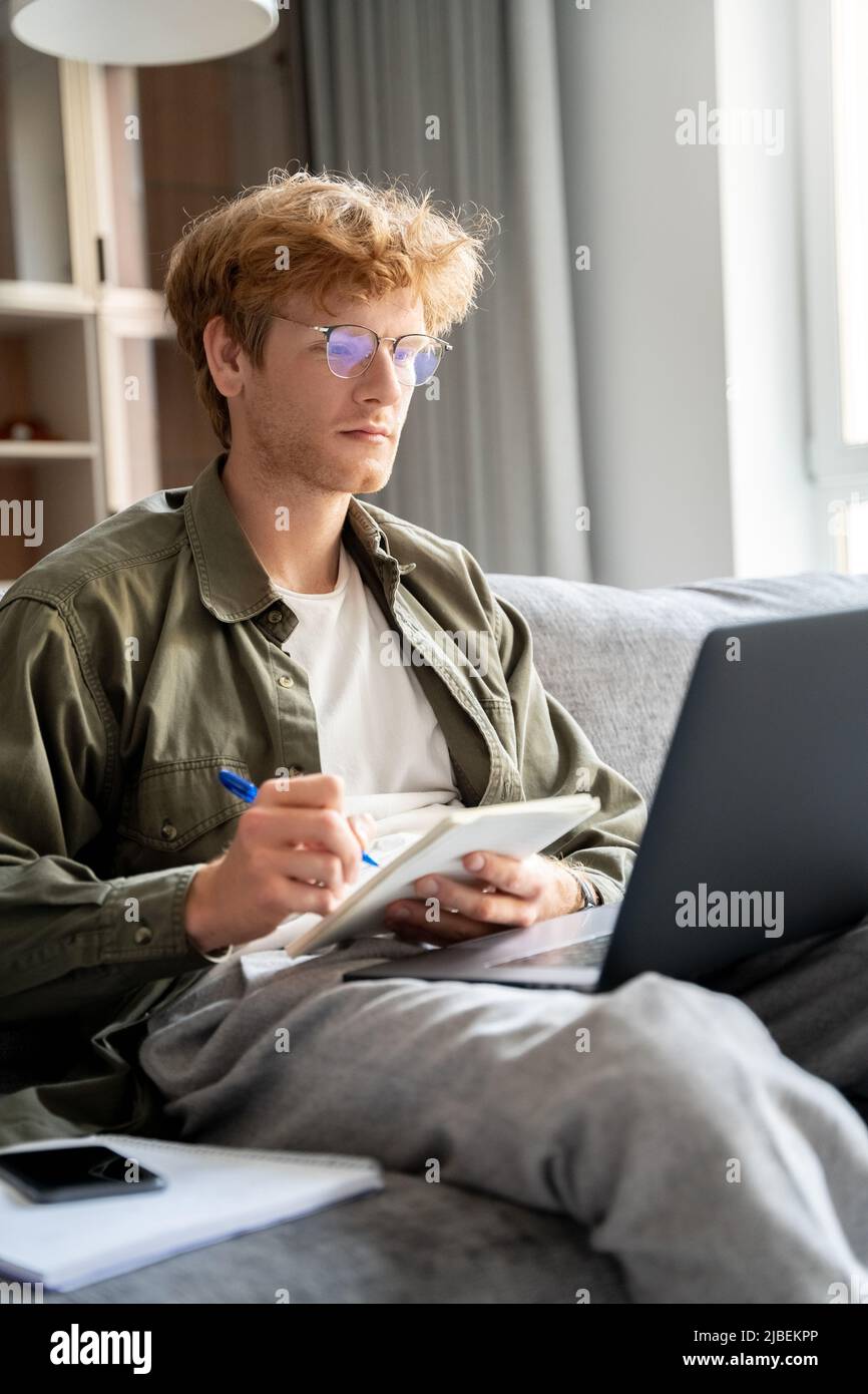 Fokussierter junger Ingwermann, der das Webinar mit einem Laptop zum Online-Lernen ansieht Stockfoto Fokussierter junger Ingwermann, der das Webinar mit einem Laptop zum Online-Lernen ansieht Stockfoto