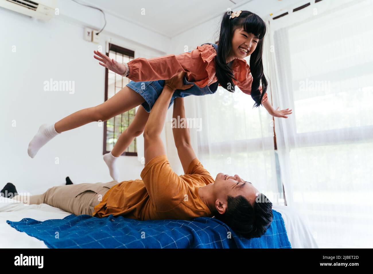 Glücklicher asiatischer Vater, der auf dem Bett liegt und die Tochter in den Armen hebt, während er zusammen im sonnenbeschienenen Schlafzimmer zu Hause spielt Stockfoto