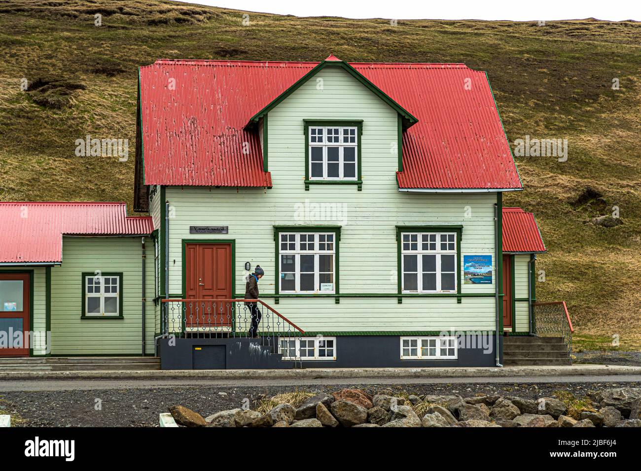 Hofsós ist ein kleines Dorf an der Ostküste von Skagafjörður. Das hellgrüne Holzhaus mit rotem Dach in Hofsós, Skagafjörður, Island, ist ein zweistöckiges Giebelgebäude mit einem schmiedeeisernen Geländer. Ein Besucher betritt gerade das Gebäude. Hofsós ist eines der ältesten Handelszentren des Landes; heute leben dort etwa 170 Menschen. Zwischen 1850 und 1914 wanderten von hier aus rund 20.000 Isländer nach Nordamerika aus. Stockfoto