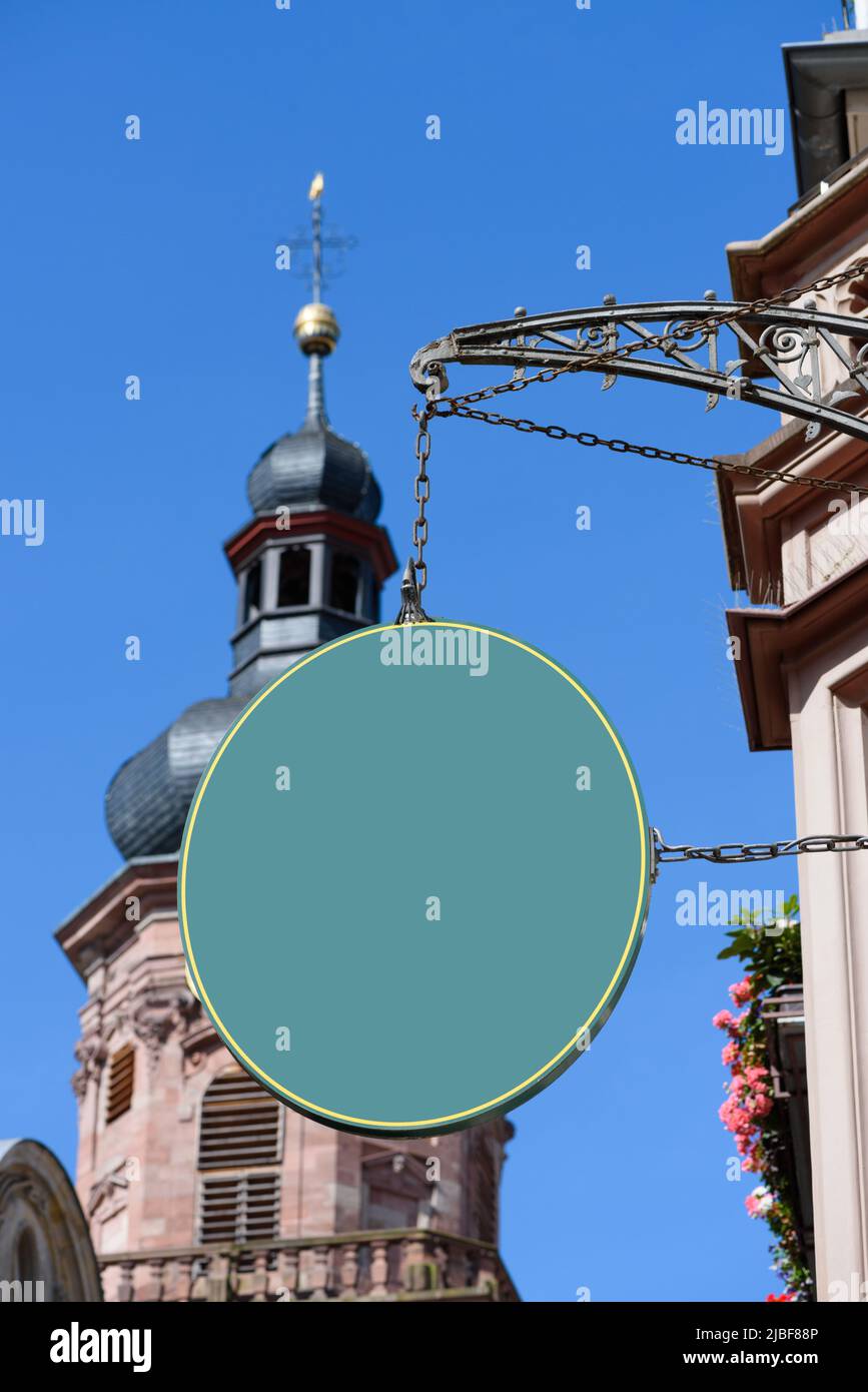 Blank grünes ovales Schild, das an einer alten Kette und einem geschmiedeten Vintage-Bogen an der Wand hängt, auf einer europäischen Straße mit Kirche im Hintergrund. Leere Plasti Stockfoto