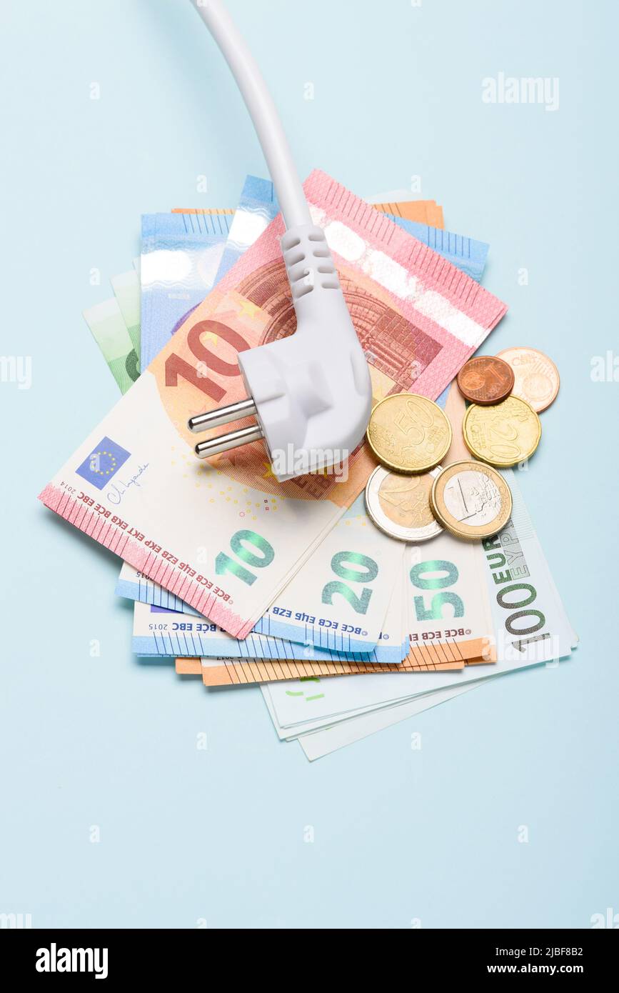 Elektrischer Netzstecker auf Euro-Banknoten und Münzen auf blauem Hintergrund. Konzept teurer Stromkosten und steigender Energiebillimpreise. Stockfoto