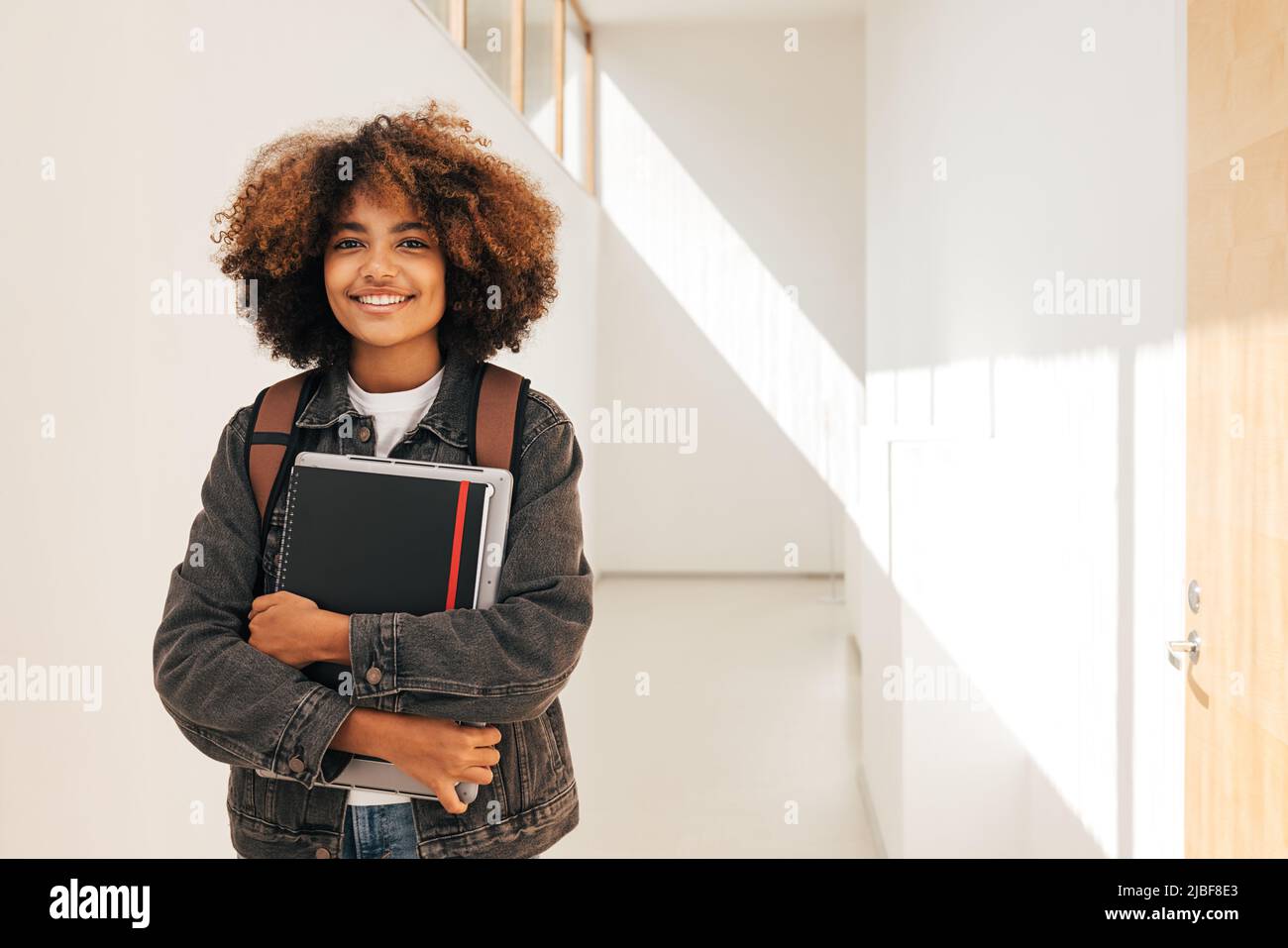 Porträt eines lächelnden Schülers, der Notebook und Laptop in der Hand hält, während er auf dem Flur steht Stockfoto
