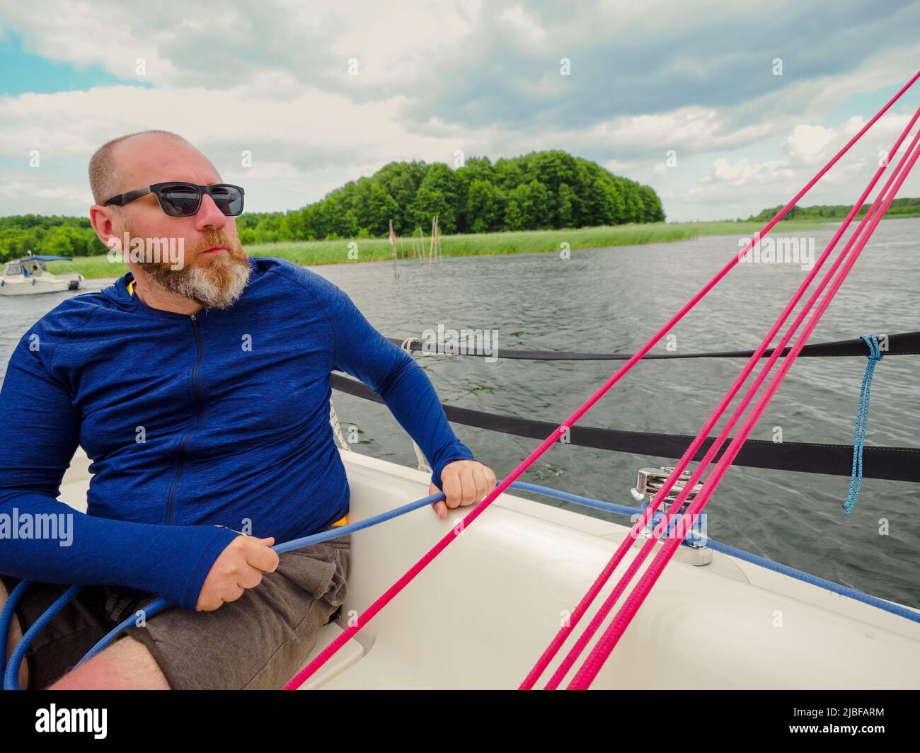 Erwachsener bärtiger Mann, der mit einer Winde ein blaues Seil auf einem Segelboot oder einer Segelyacht zieht Stockfoto