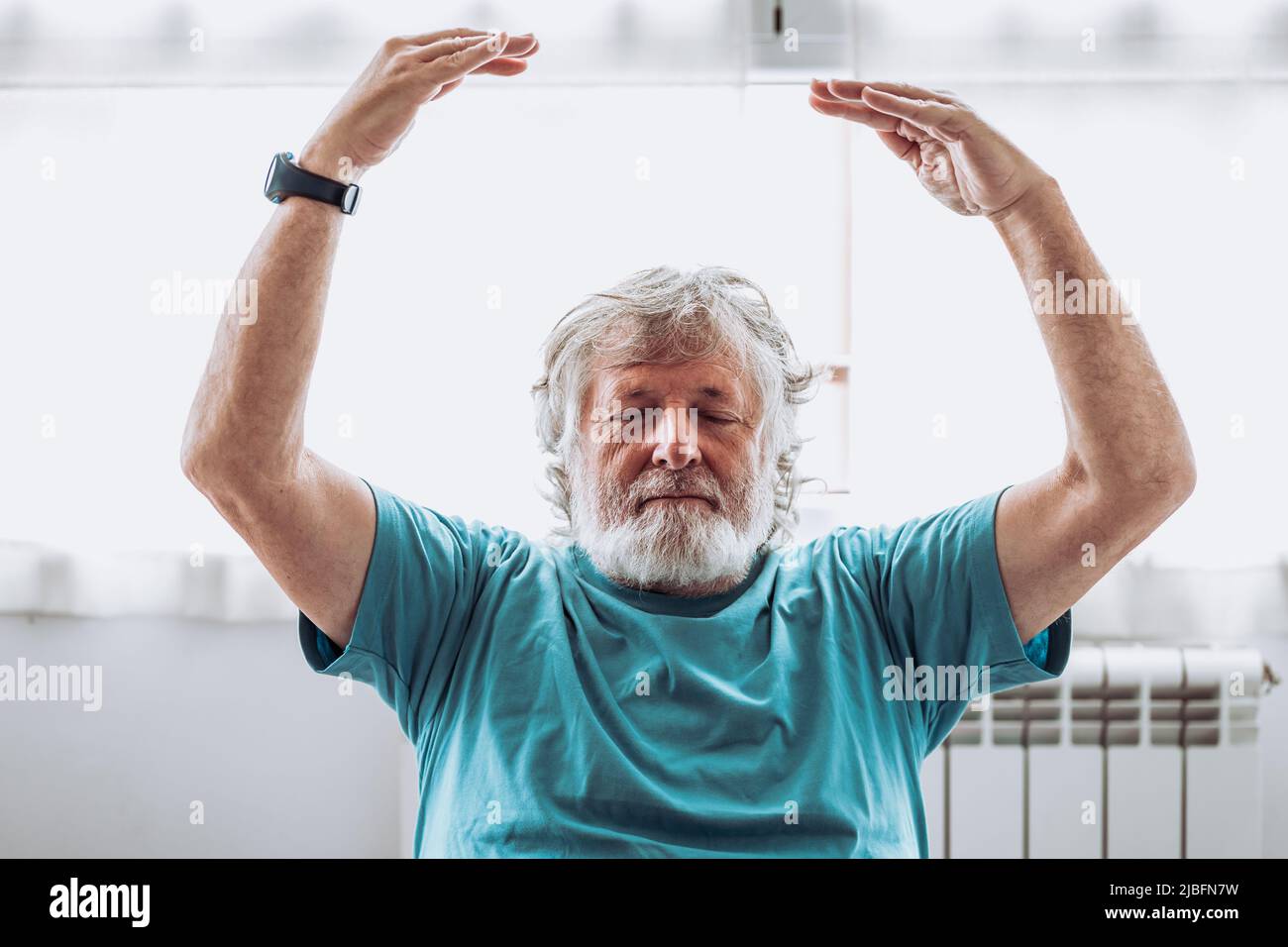 Älterer Mann im blauen T-Shirt, der die Arme hebt und mit geschlossenen Augen gegen das Fenster meditiert, während der Yoga-Sitzung am Morgen zu Hause Stockfoto