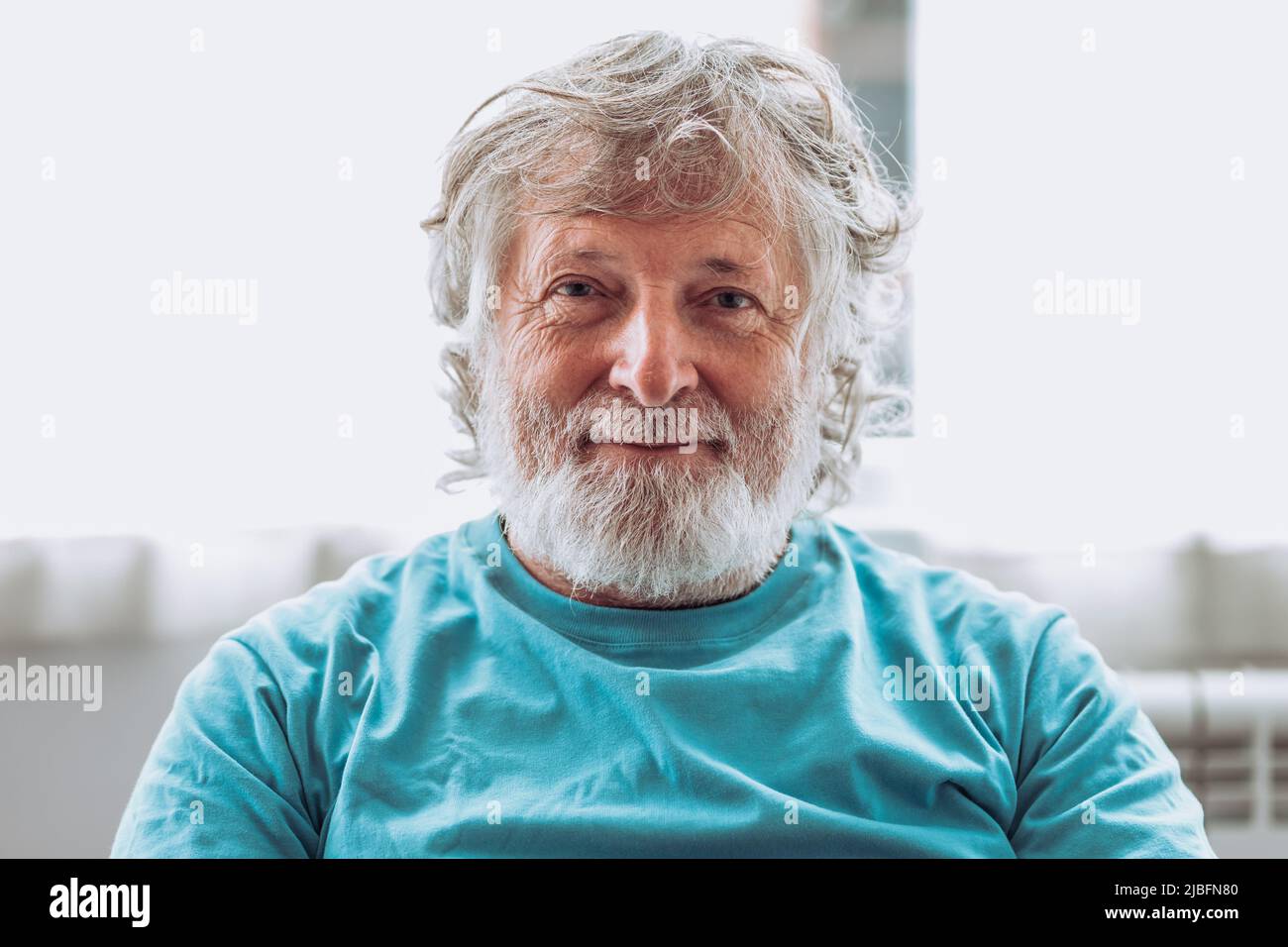 Älterer Mann mit grauem Bart vor verschwommenem Hintergrund und Blick auf die Kamera zu Hause Stockfoto