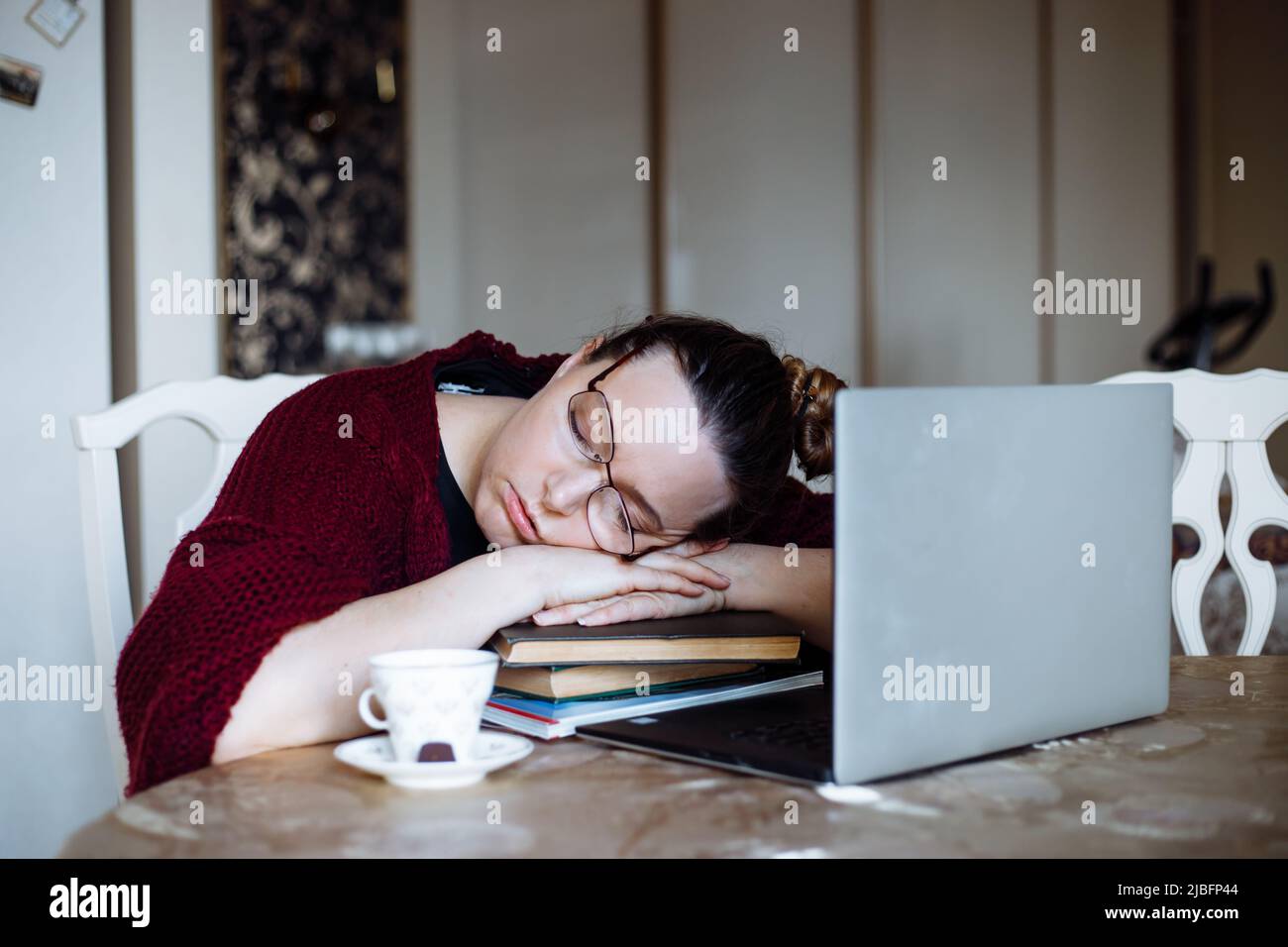 Porträt einer müden Frau mittleren Alters, die am Tisch in der Nähe des Laptops saß und auf gekreuzten Armen auf Bücherhaufen schlief, saß zu Hause. Stockfoto